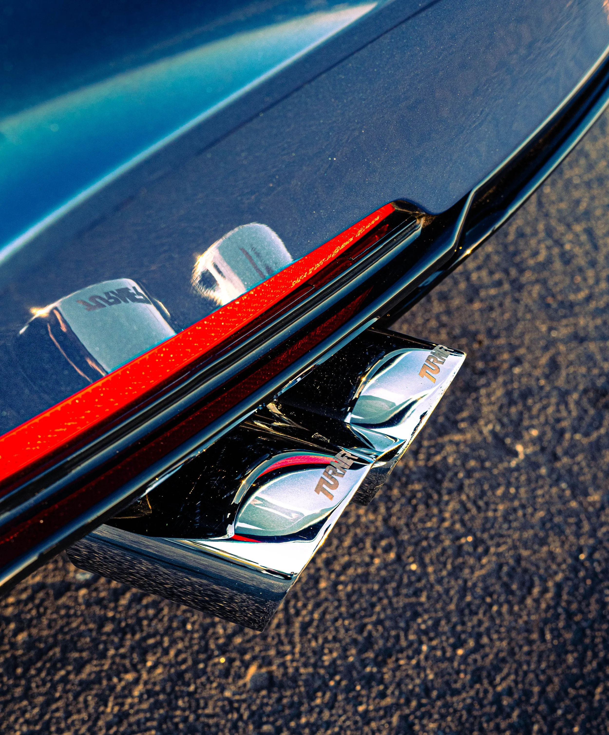 Close-up of a shiny, dark-colored car with a red stripe and a 'TURBO' badge, parked on a textured asphalt surface with sunlight reflecting off the surface.
