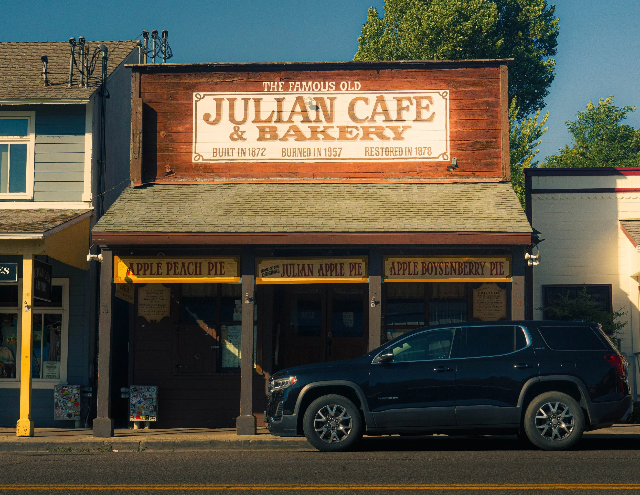 Historic brick building with a wooden sign that reads 'Julian Cafe & Bakery,' built in 1872, burned in 1957, restored in 1978. The building has a storefront with signs advertising apple pies and other baked goods, and a car parked in front.