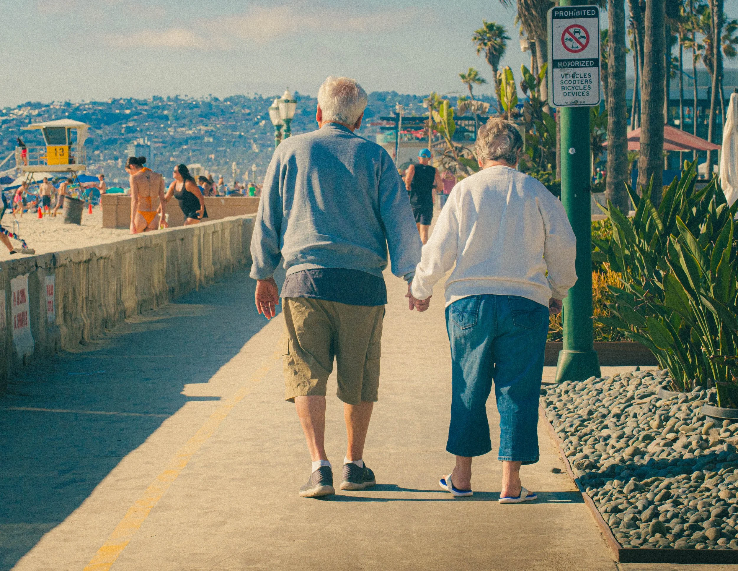 Two elderly people, one man and one woman, walking hand in hand on a paved seaside pathway. The beach is crowded with people, umbrellas, and a lifeguard tower in the background. There are palm trees and a sign indicating prohibited vehicles, scooters