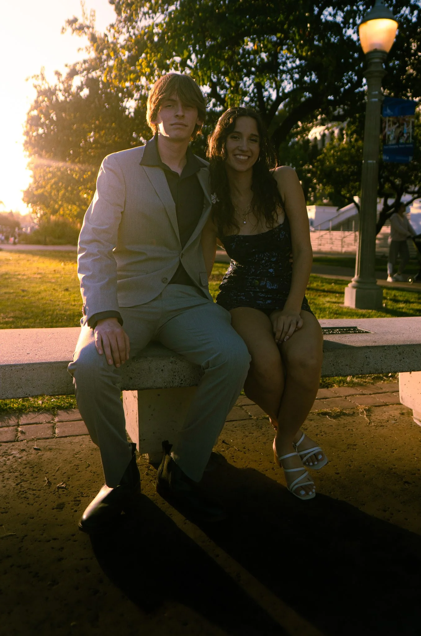 A young man wearing a grey suit and black shirt sitting on a concrete bench beside a young woman in a black dress with floral pattern, smiling, in a park during sunset with trees and a lamppost in the background.