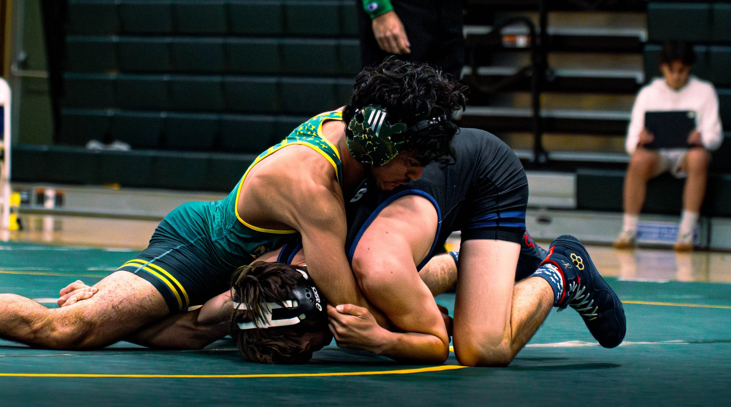 Two wrestlers on the mat, one in a yellow and black singlet and the other in a black singlet, engaged in a wrestling match with a referee in the background.