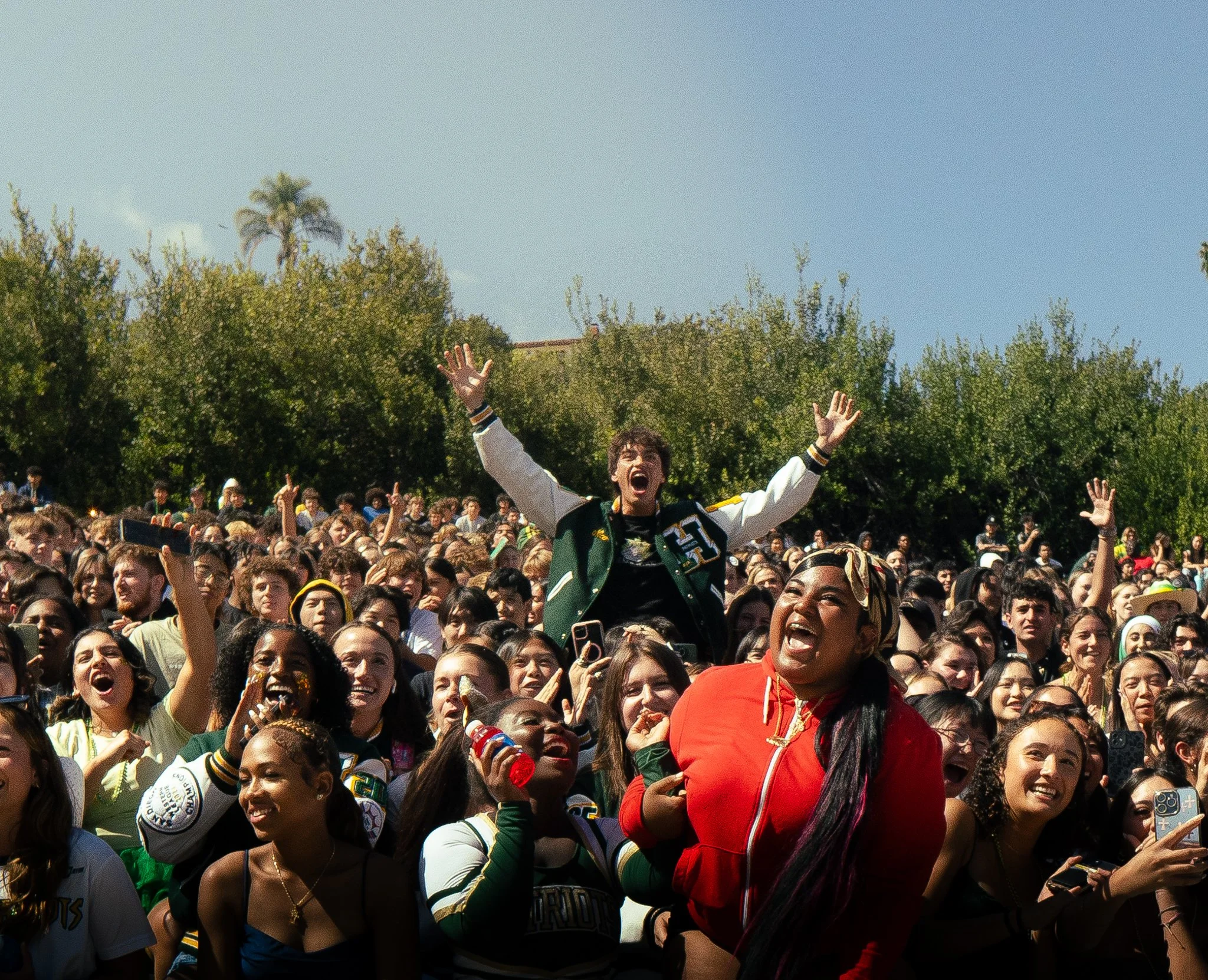 Crowd of diverse smiling people outdoors under clear blue sky, celebrating, with a woman in a red jacket smiling in the foreground.