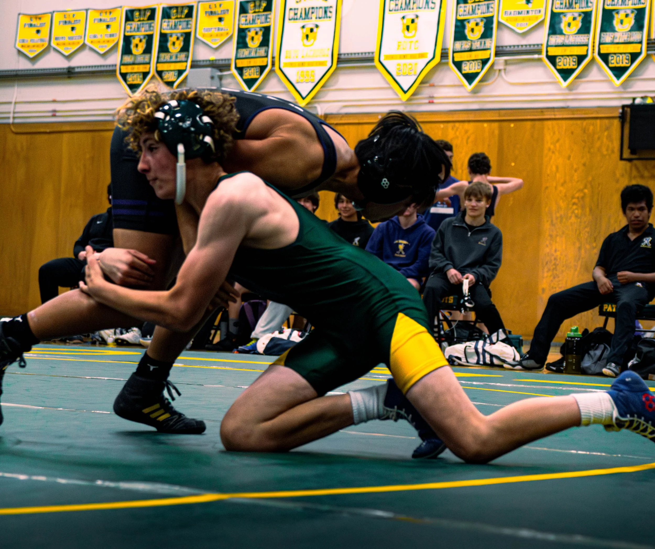 Two wrestlers competing on a mat in a gymnasium, with spectators and gym banners hanging in the background.