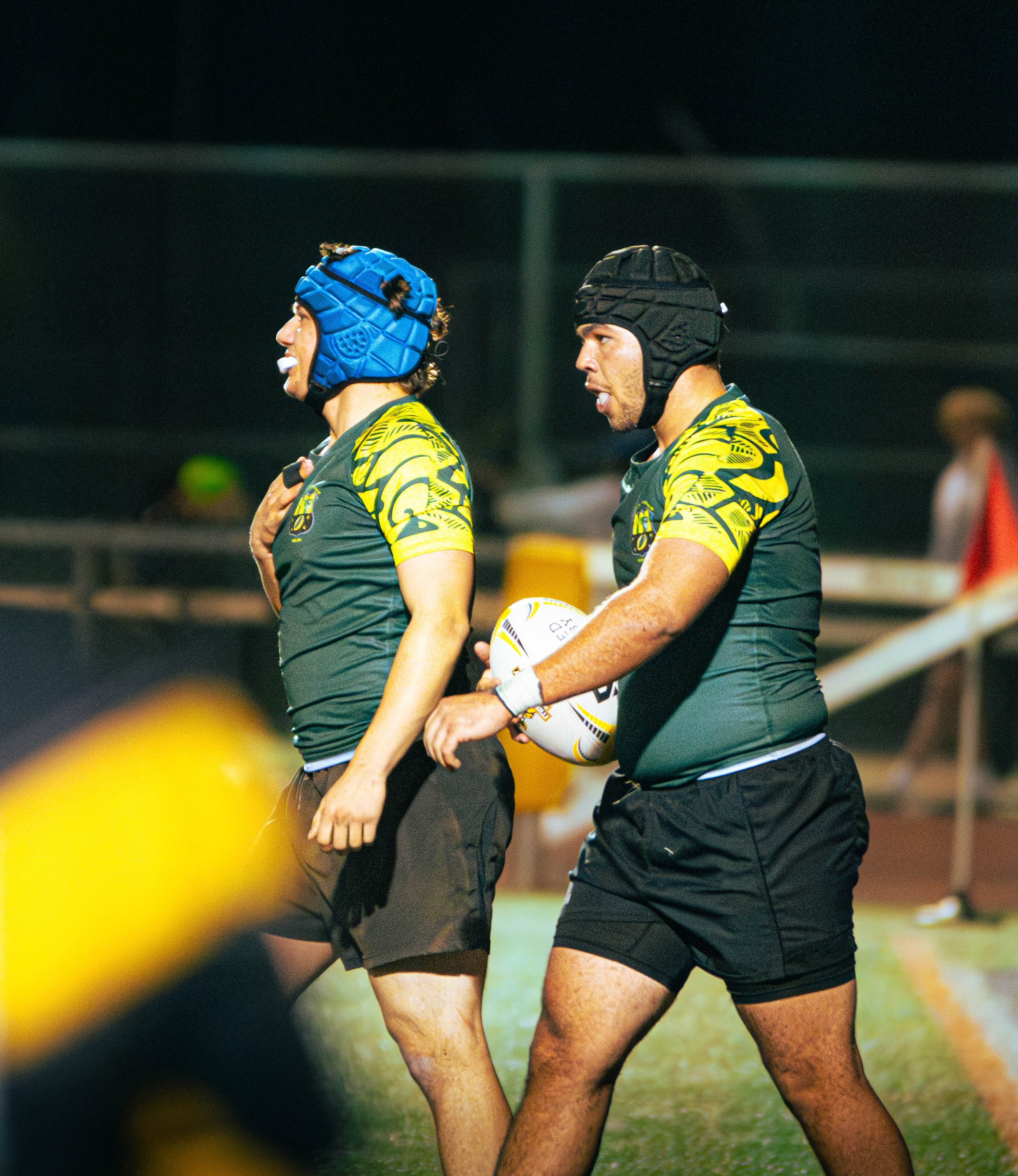 Two rugby players wearing black and yellow jerseys and protective headgear, one with a blue helmet and the other with a black helmet, are standing on a rugby field during a game at night, with blurred spectators in the background.
