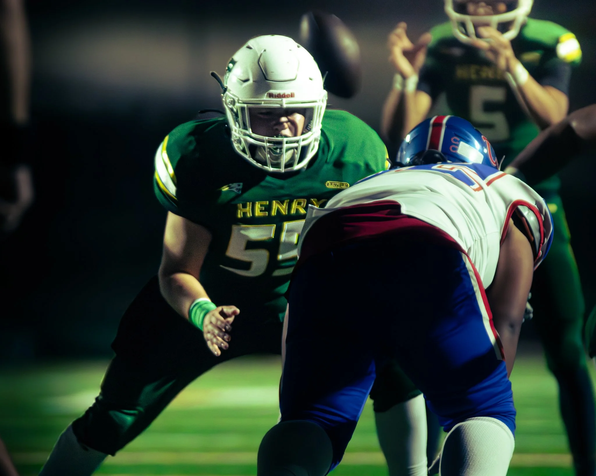 A football game with players in green and black uniforms, one player wearing a helmet with a face guard, engaging in a tackle or tackle attempt.