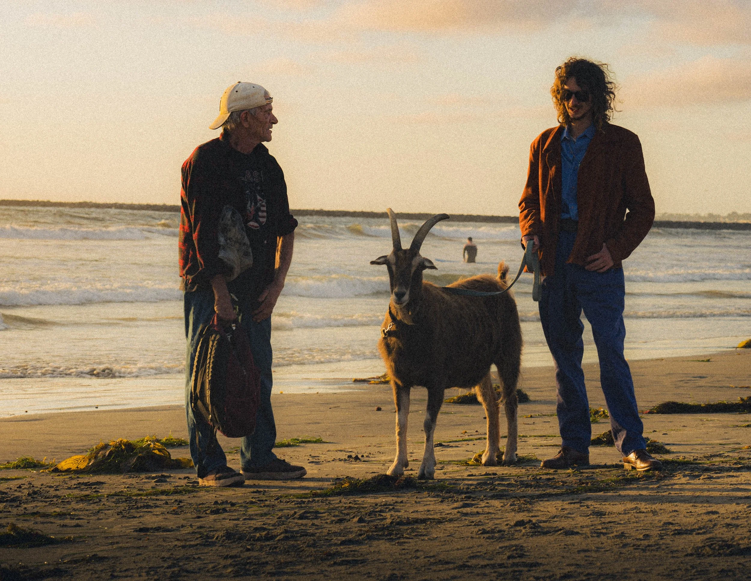 Two men and a goat on a beach during sunset, one man in a baseball cap and sweatshirt, the other with long curly hair wearing sunglasses and a brown jacket, both standing near the water's edge.
