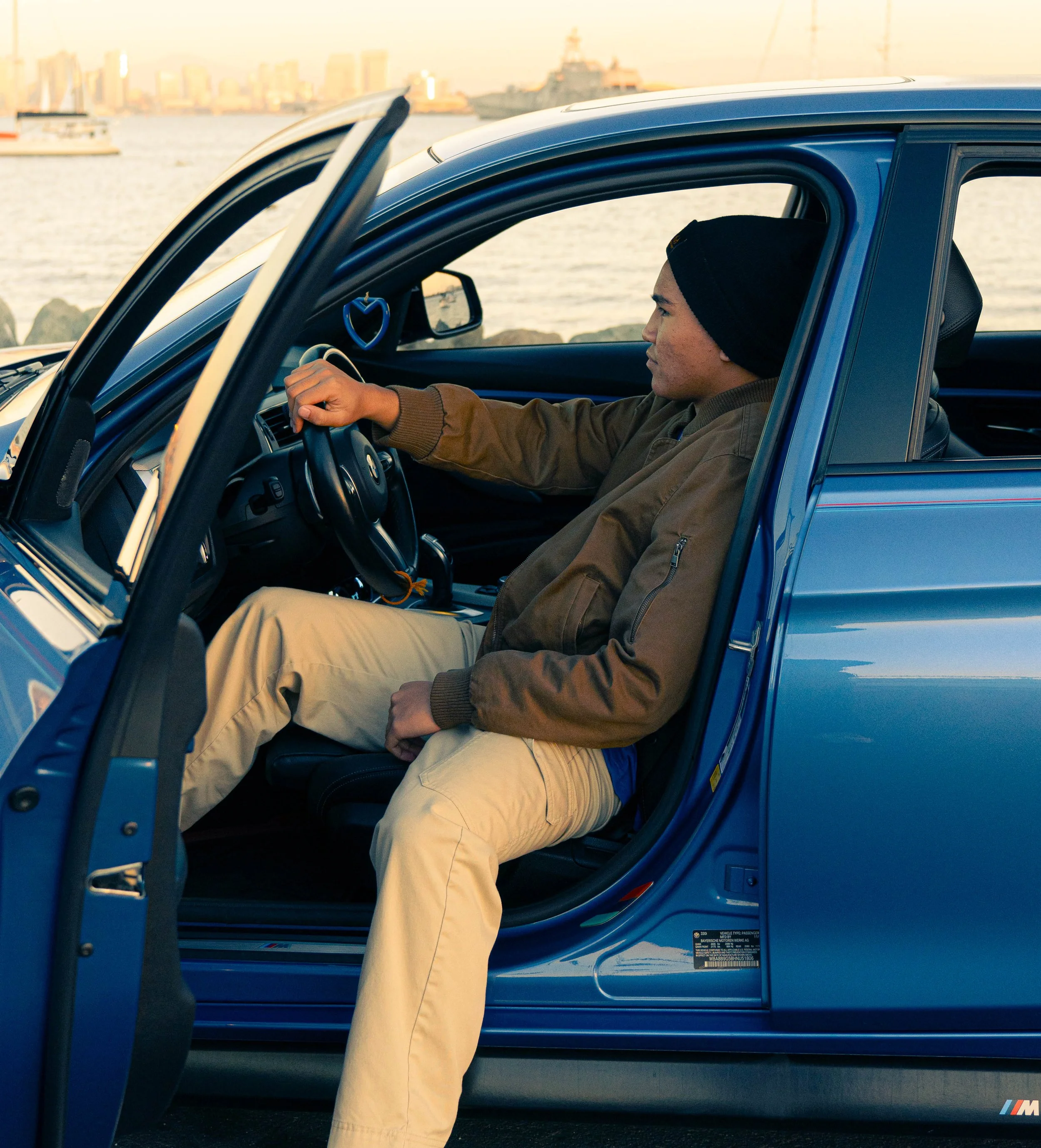A man wearing beige pants, a brown jacket, and a black beanie is sitting in a blue car with the door open, by the water with boats and a city skyline in the background during sunset.