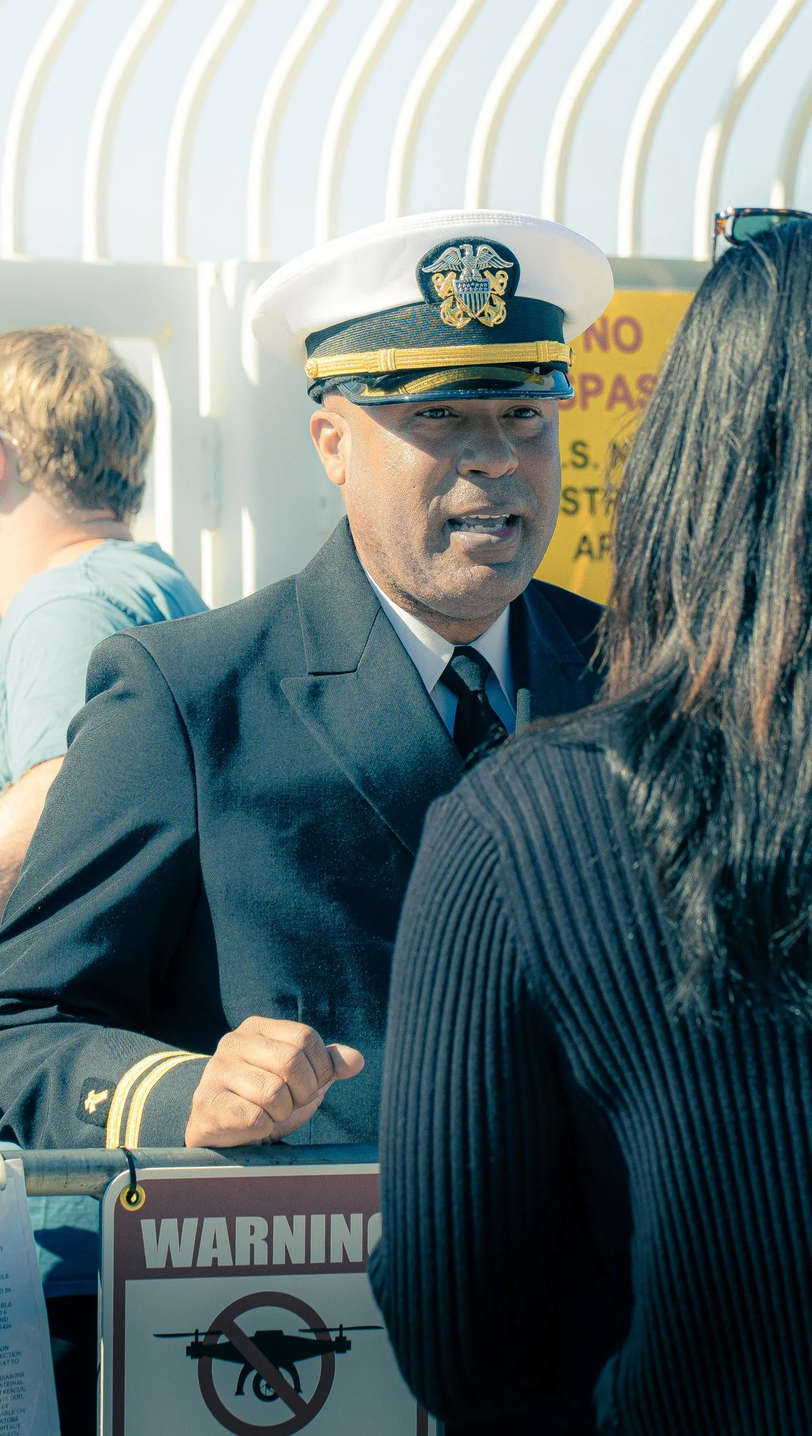 A man in military uniform and a white cap speaking to a woman with long dark hair at a protest or rally, with a warning sign in the background.