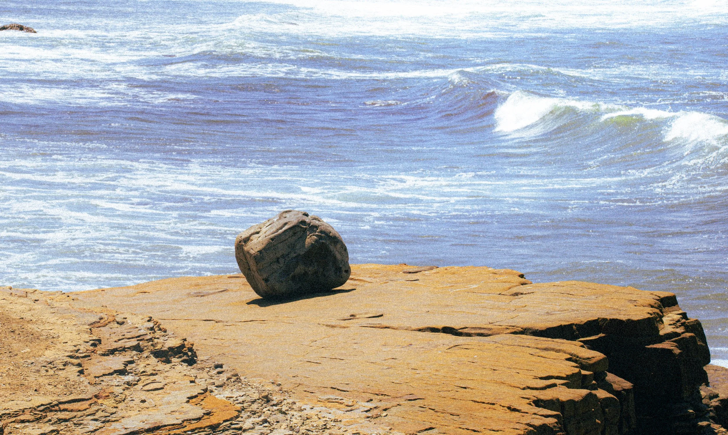 A large rock on a sandy beach with ocean waves in the background.