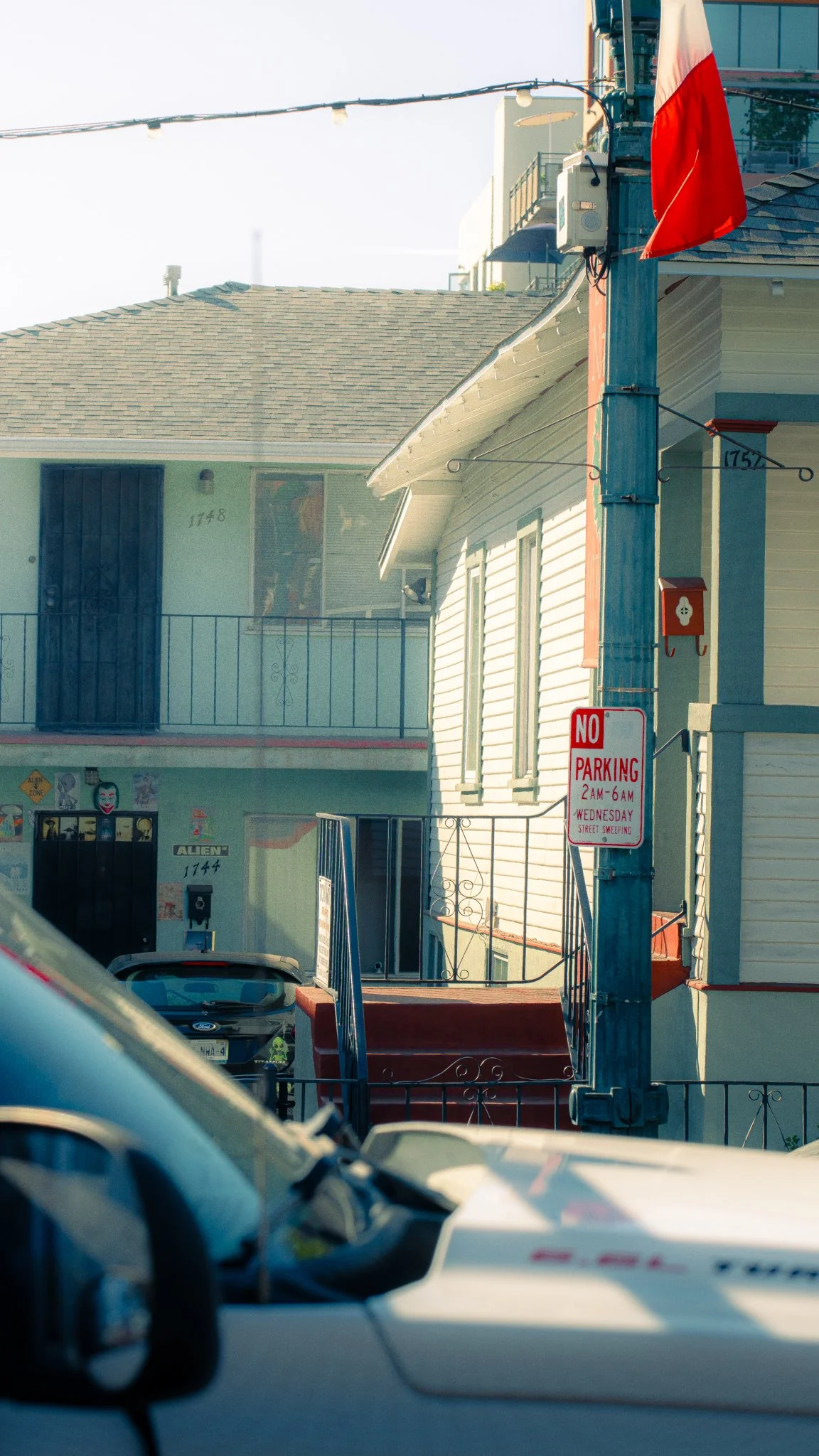 A street scene with houses, a pole with street signs, a red and white flag, and wires overhead. Several parked cars are visible in the foreground.