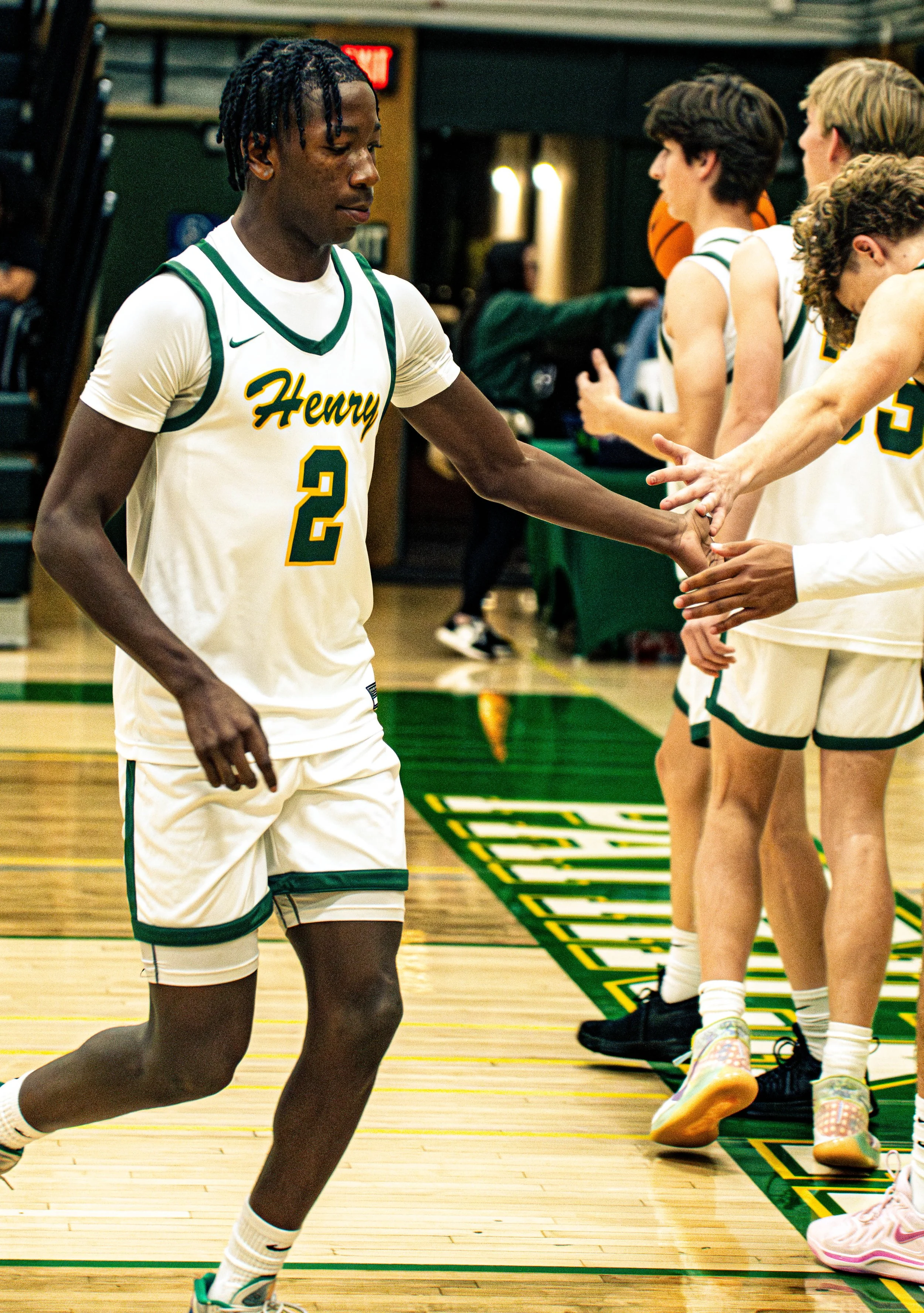 A high school basketball team (Henry) warming up before a game in the gymnasium. Players are in their white and green uniforms. One player is running, and others are standing near the sideline.