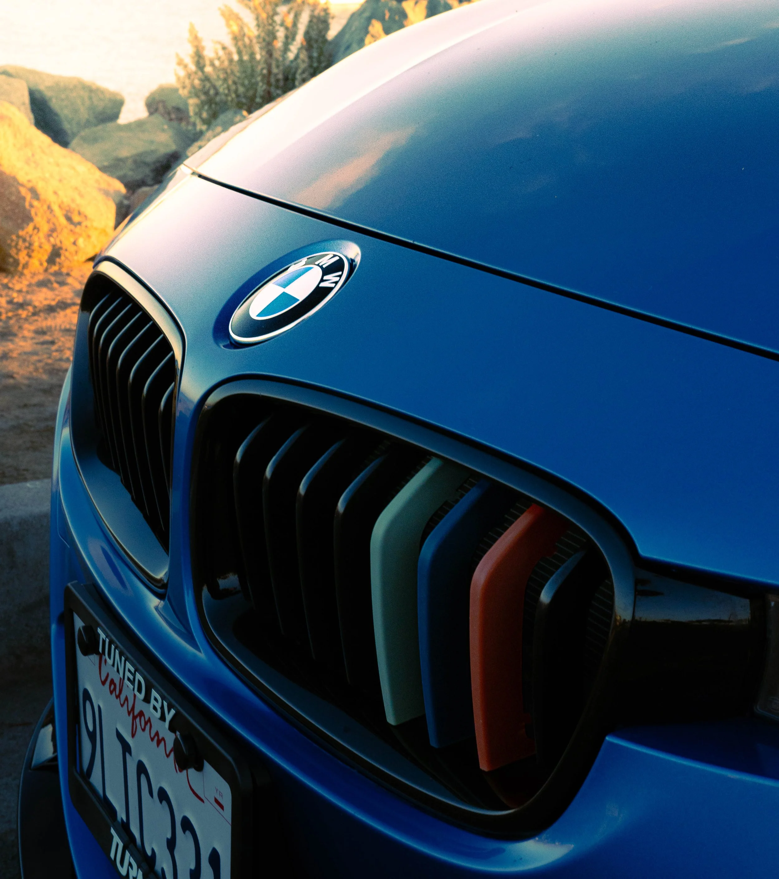 Close-up of front part of a dark blue BMW car with the BMW logo and a black grille, with a background of rocks and trees at sunset.
