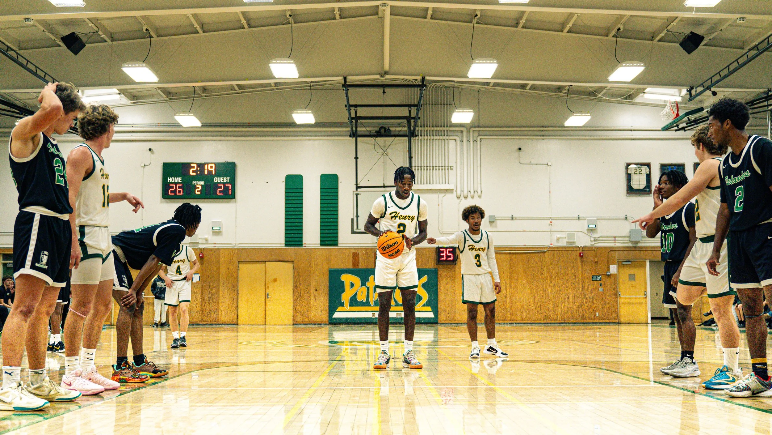 A basketball team gathering on a gym court, with players standing on both sides of the court, facing inward and listening to their coach during a game or practice. The scoreboard shows a score of 28-27 with 2 minutes and 19 seconds remaining in the s
