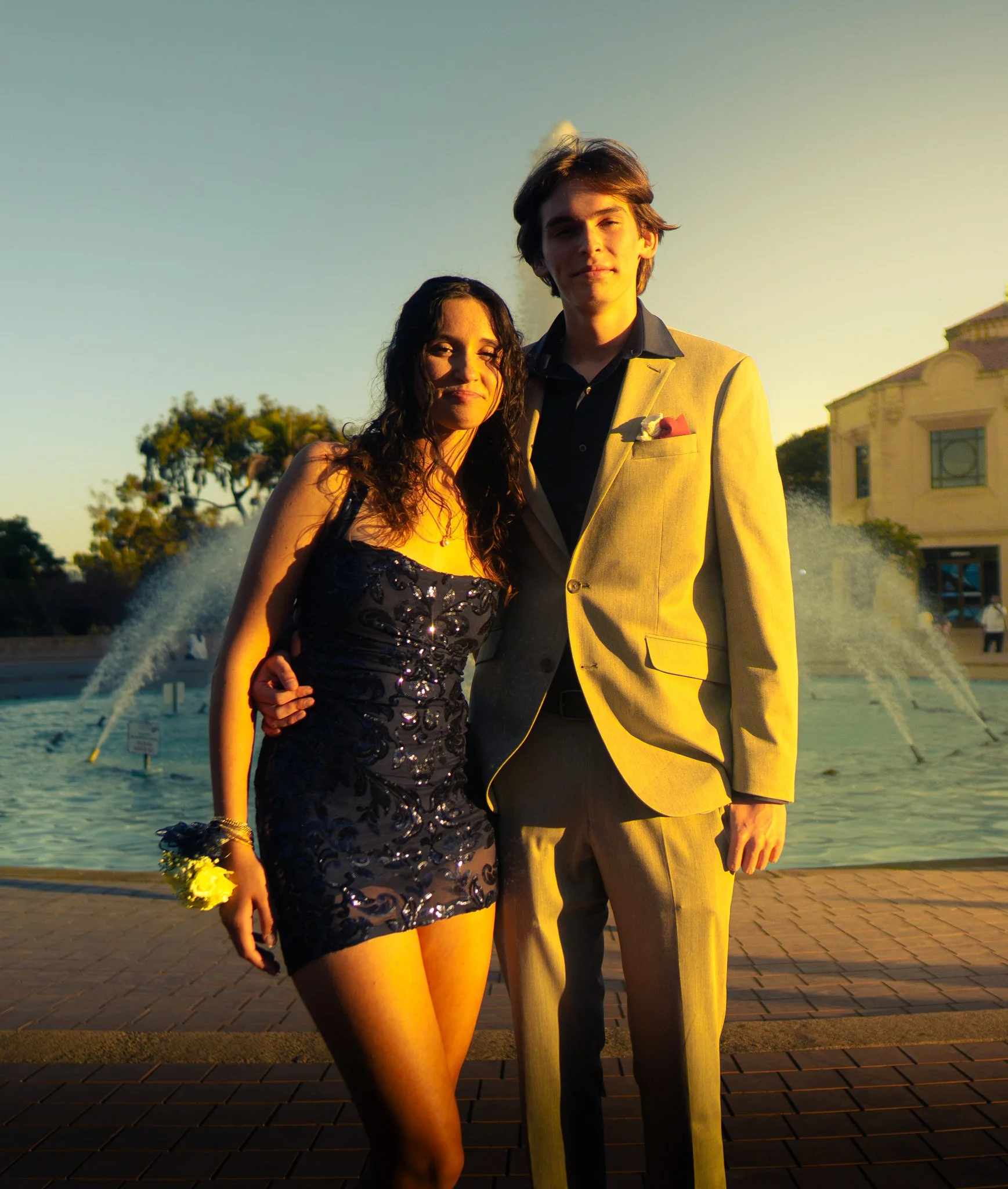 A young couple stands close together outdoors during sunset, with water fountains in the background. The woman wears a black, sequined dress and holds a bouquet of yellow flowers, while the man wears a light-colored suit jacket with a boutonniere.