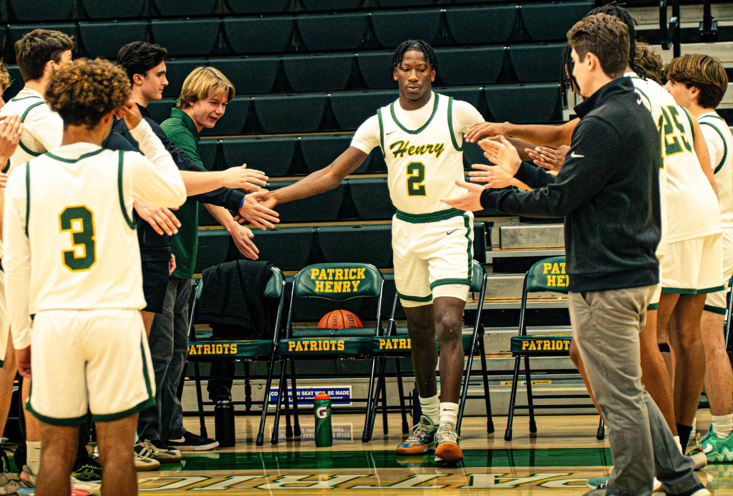 A basketball team in white and green uniforms gathered in a gym, with one player in the center dribbling and others standing around, some clapping and others reaching out for high-fives.