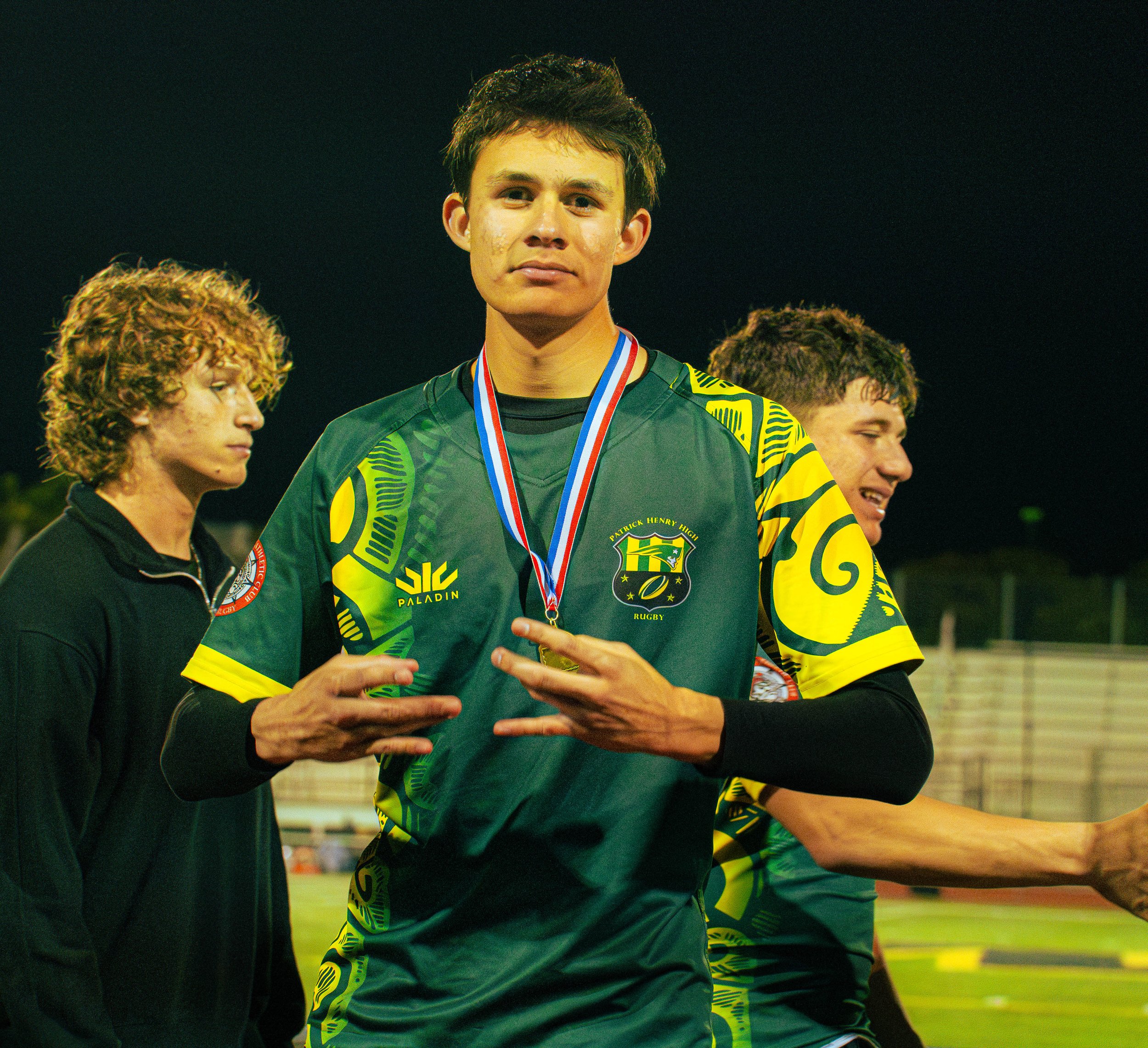 A young male athlete in a green and yellow sports jersey with a medal around his neck, standing on a sports field at night. He is flanked by two other young men, both smiling and wearing similar jerseys, under stadium lights.