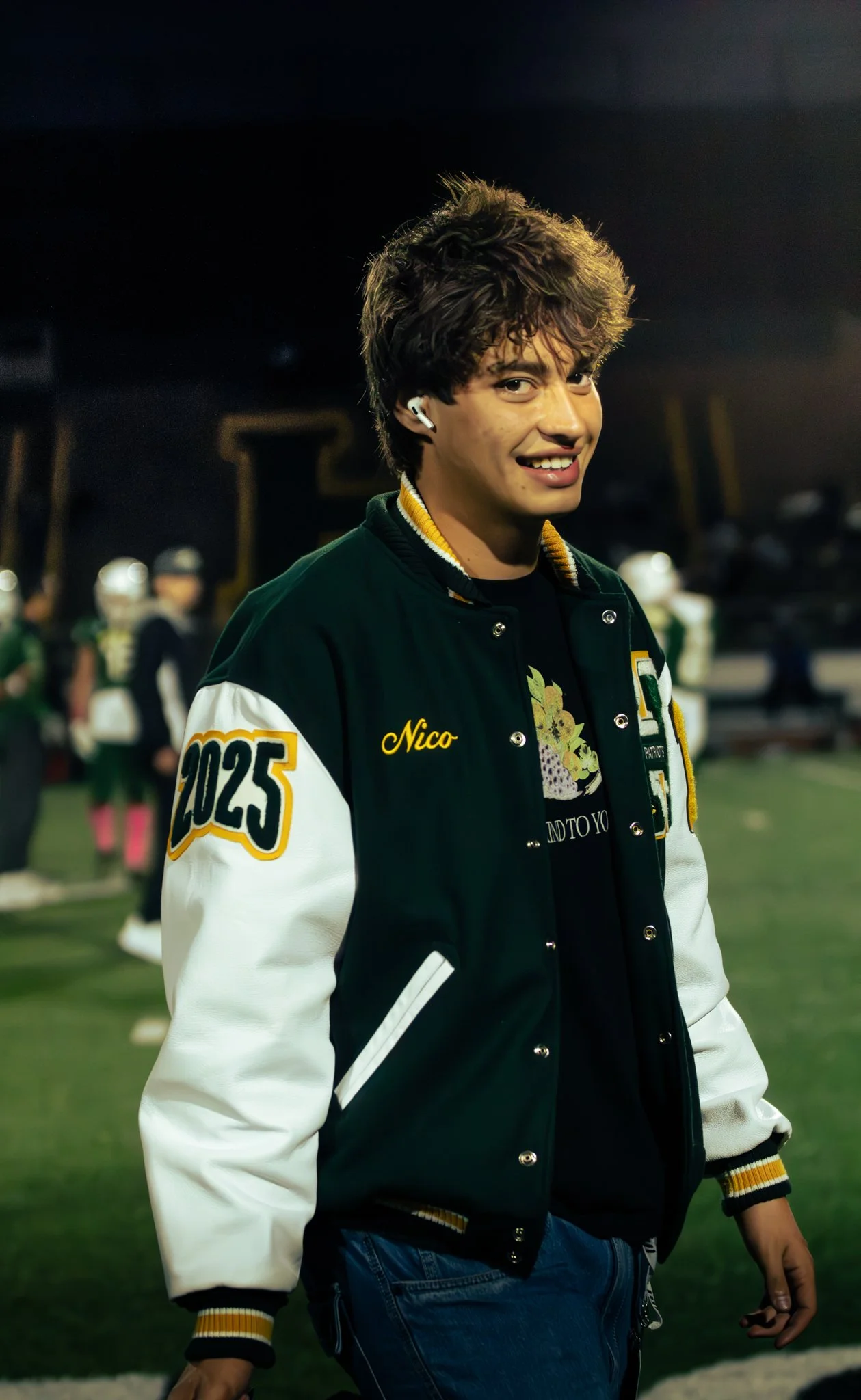 Teenage boy with curly hair wearing AirPods, a black and white varsity jacket with yellow accents and the name 'Nico' embroidered on it, standing on a sports field at night, smiling.