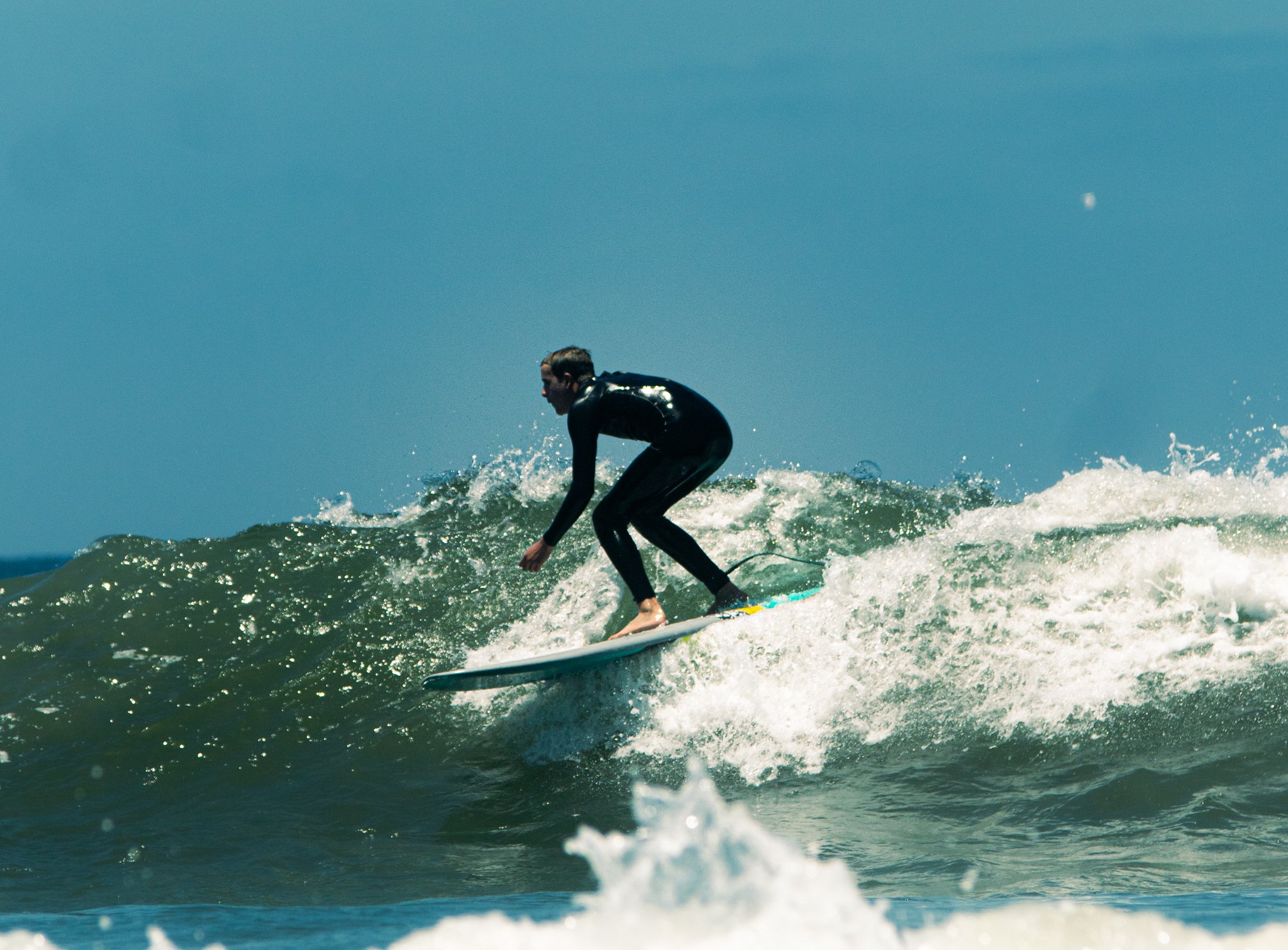 A person wearing a wetsuit surfing on a wave under a clear blue sky with a visible moon.
