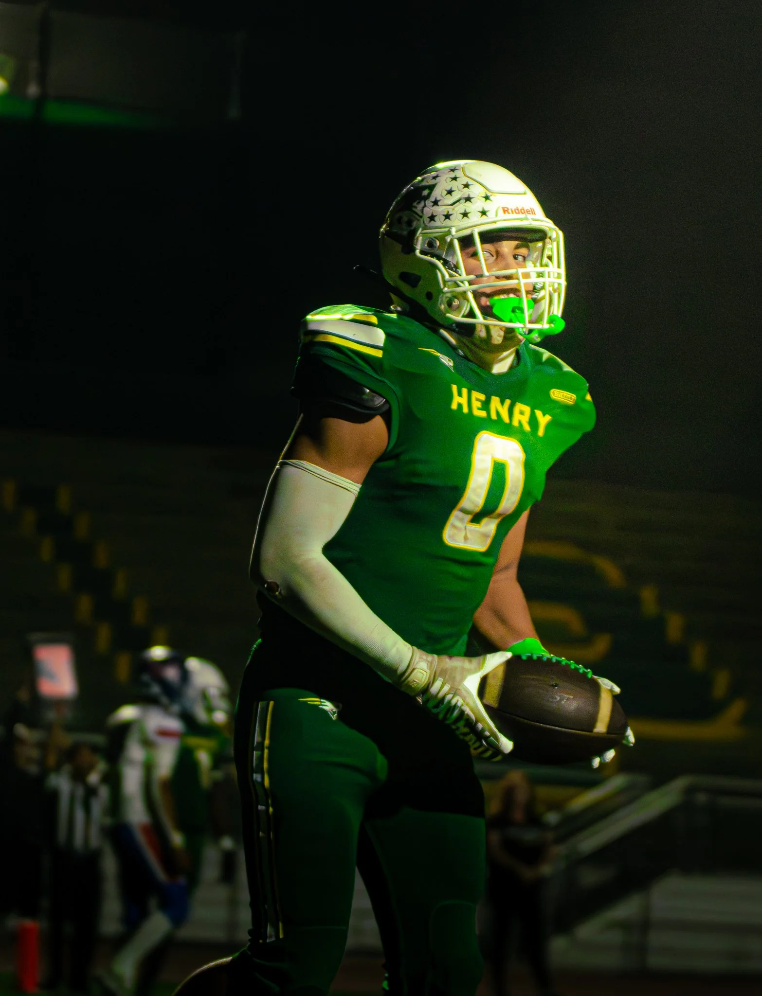 Football player wearing green and yellow uniform with the name 'Henry' and number 0, wearing a helmet with stars, holding a helmet, and standing on a field at night.