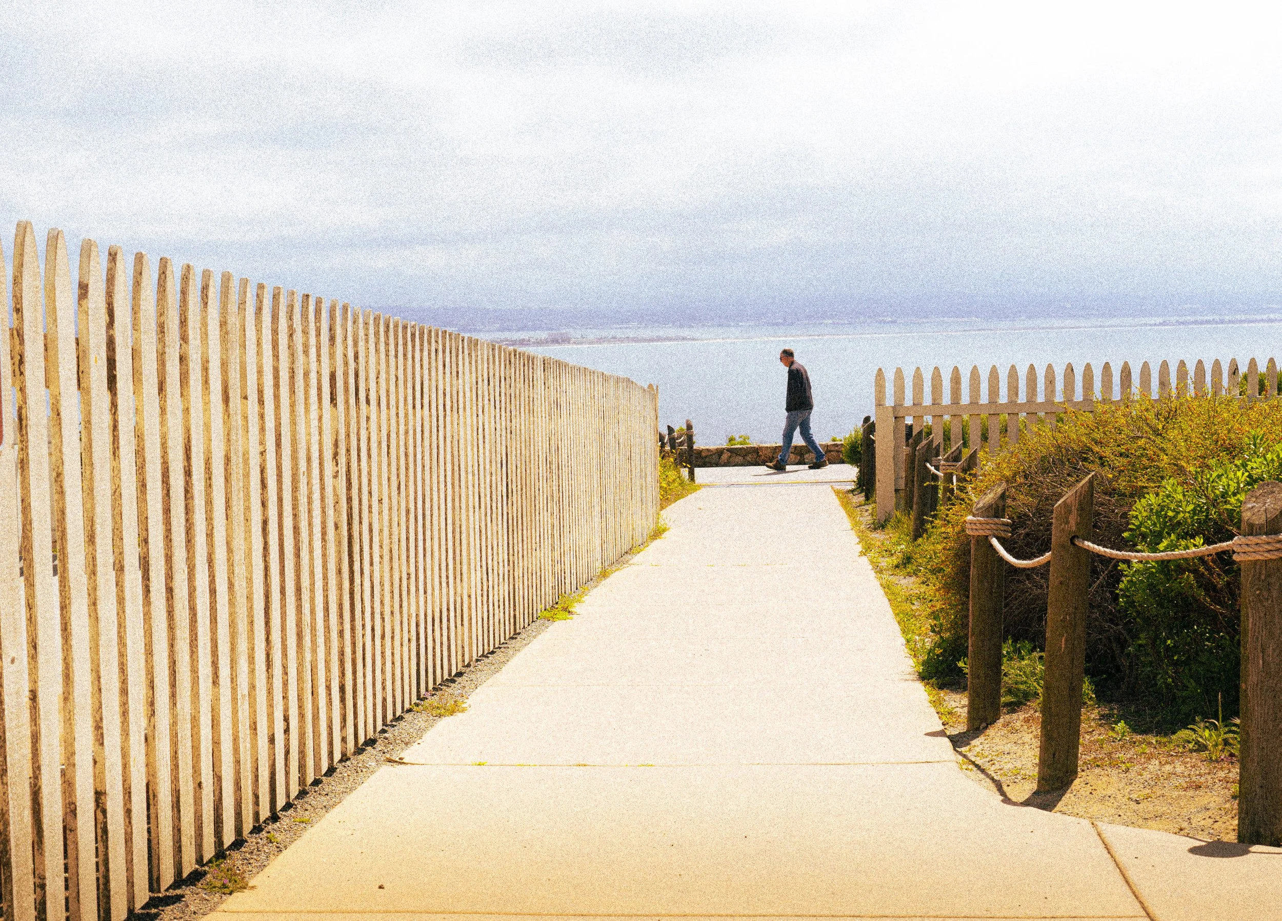 A man walking along a paved path with a wooden fence on both sides, overlooking a body of water with a distant shoreline under a partly cloudy sky.