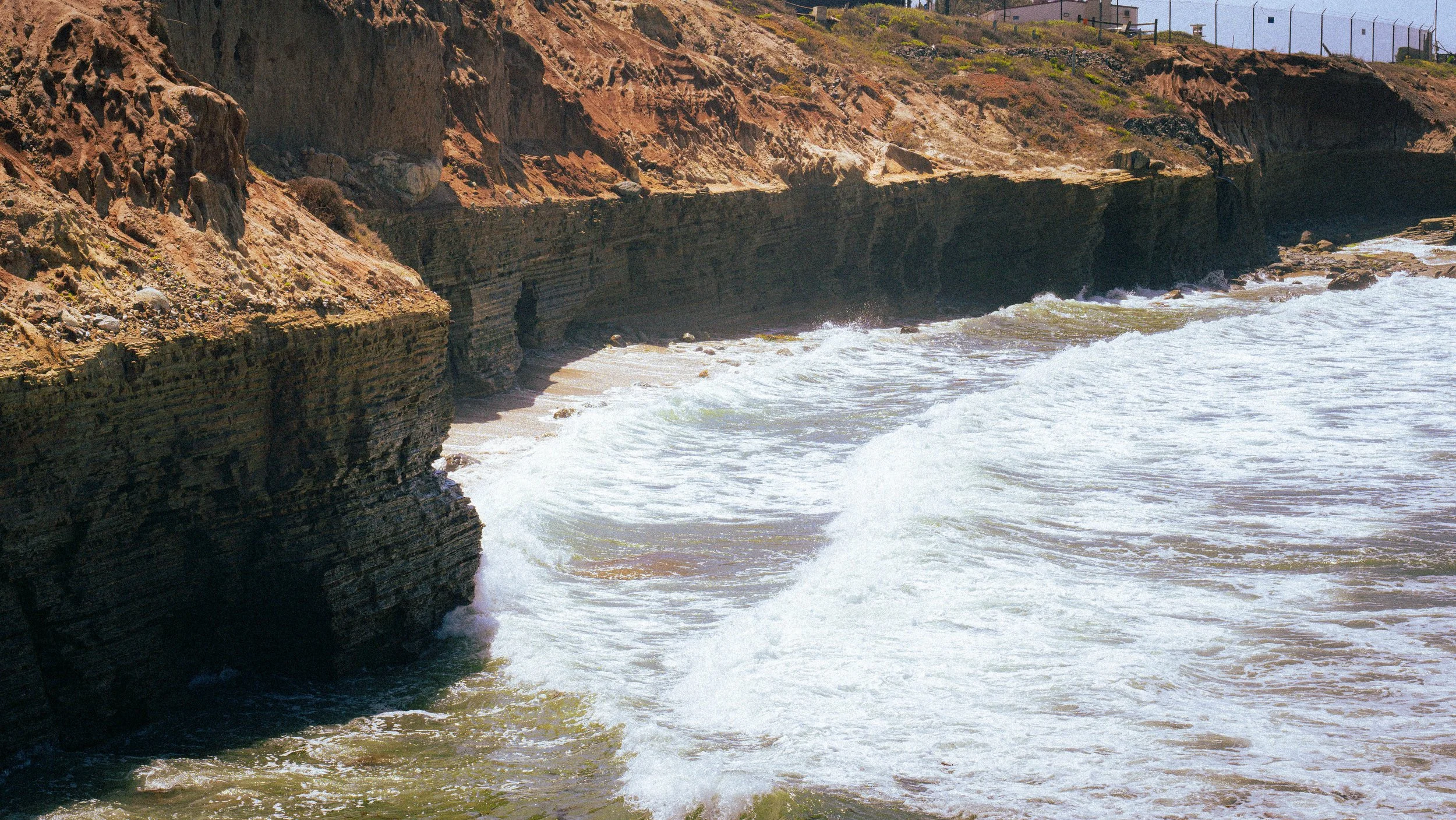 Cliffs overlooking the ocean with waves crashing against the rocks and a walkway with a railing at the top of the cliffs.