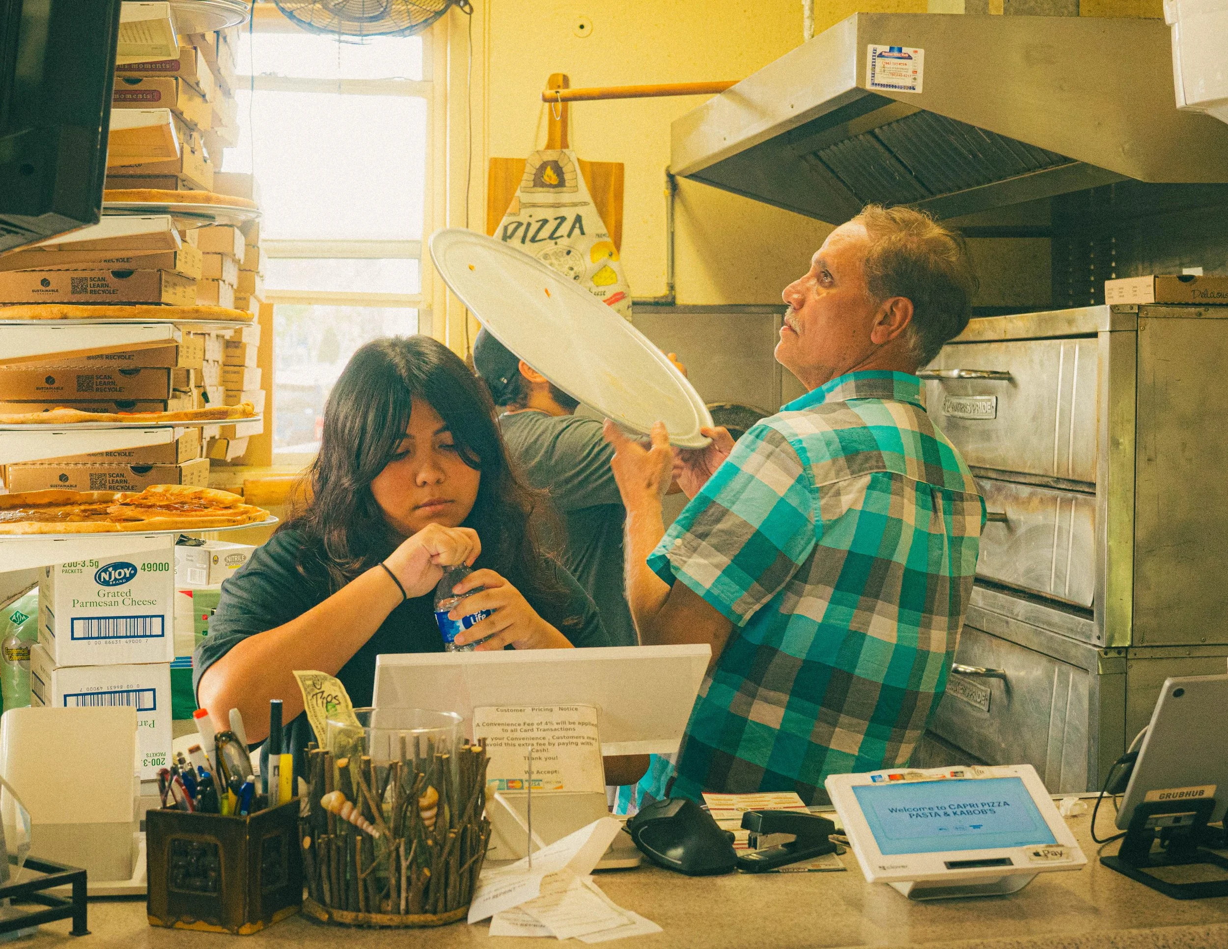Inside a pizza restaurant with a counter, pizza boxes stacked on the shelves, a woman with dark hair picking pizza slices, and a man in a blue checkered shirt holding a pizza tray. There are menus, a tablet, and various supplies on the counter.