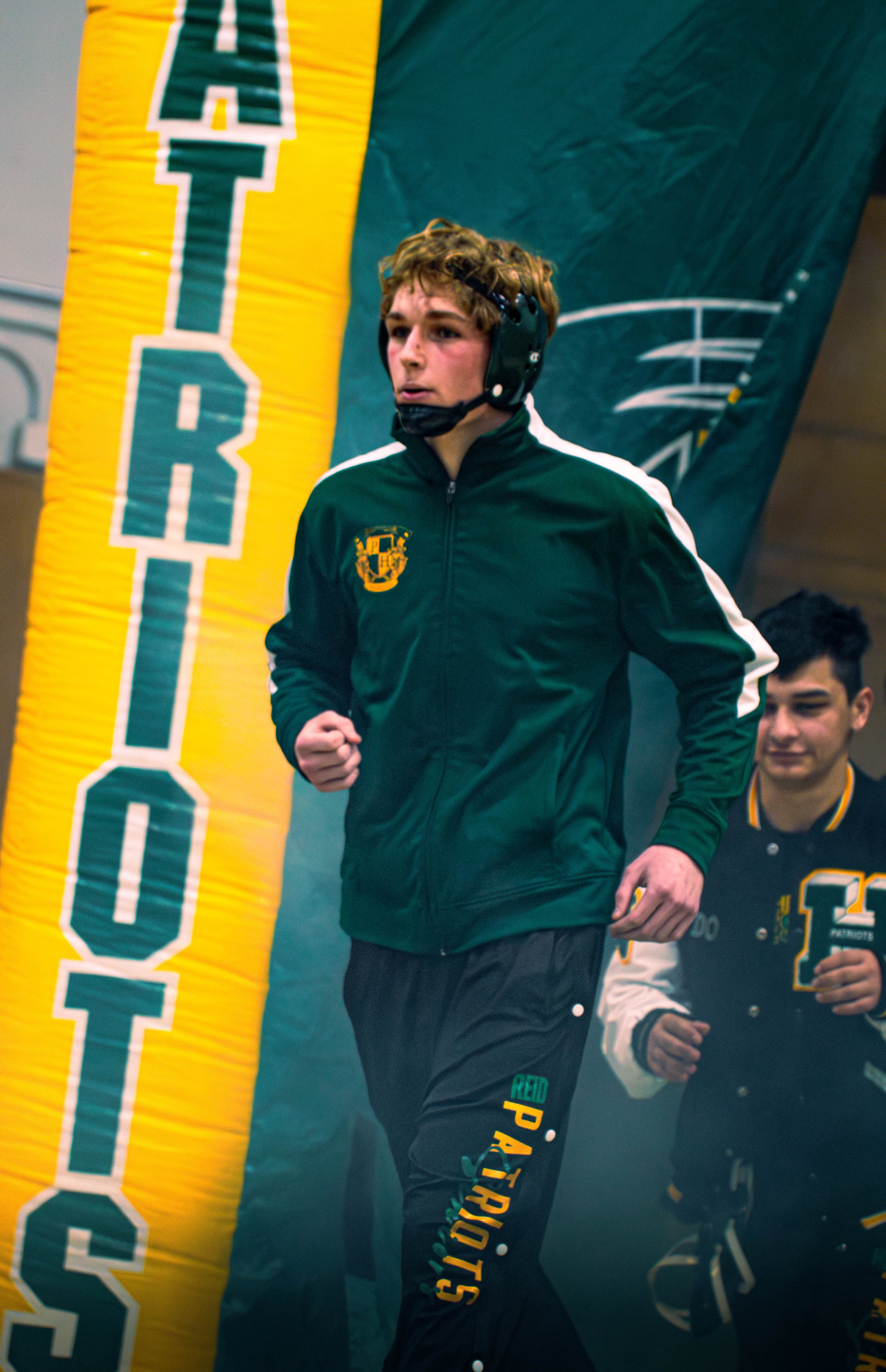 A young man running in sportswear, wearing a green and yellow jacket with a crest on the chest and black pants with colorful text on the side. Two other young men are in the background, one smiling.