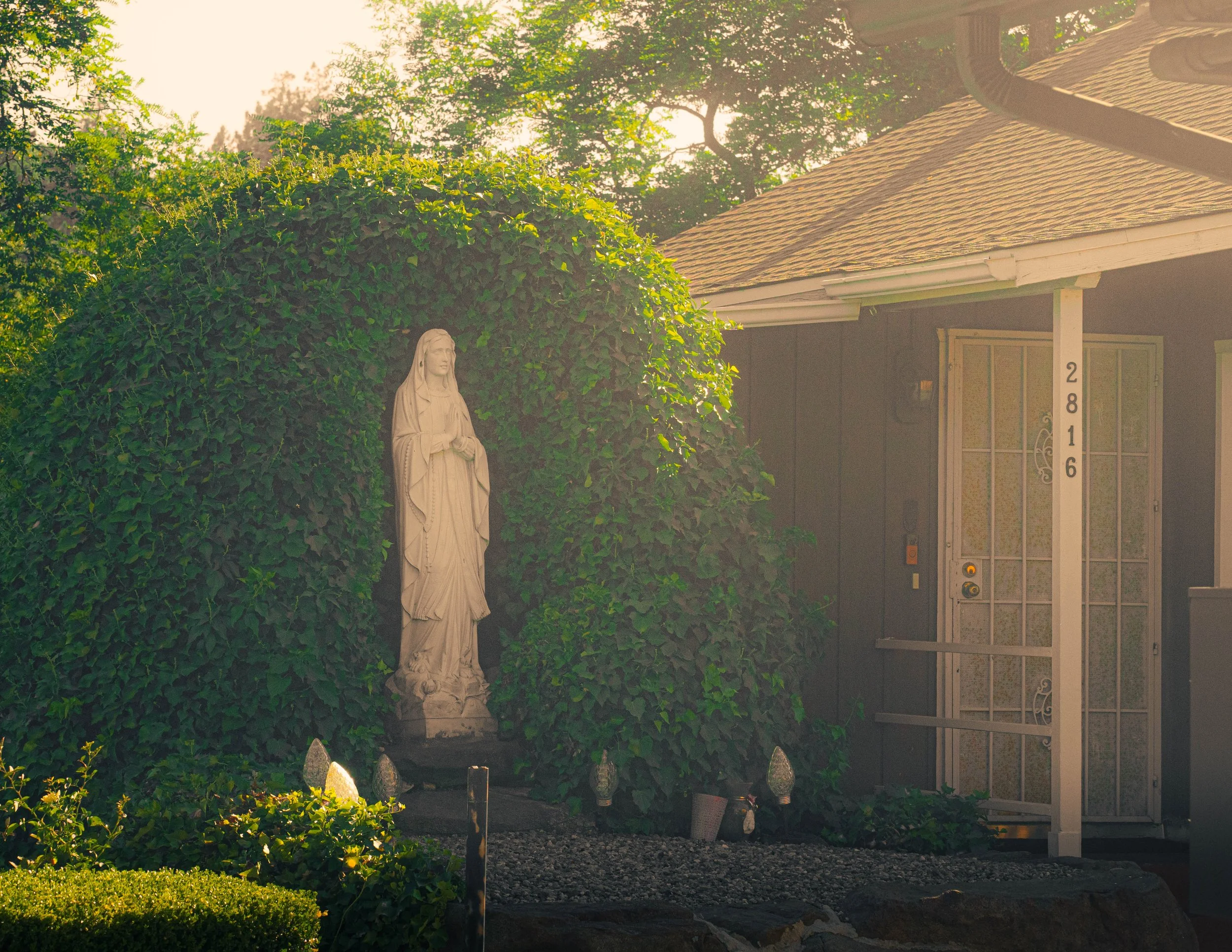 A religious statue of Jesus Christ standing in a garden next to a bush. Behind it is a house with a shingle roof and an American-style porch. The house has a white pole with house numbers. The scene is bathed in warm sunlight.