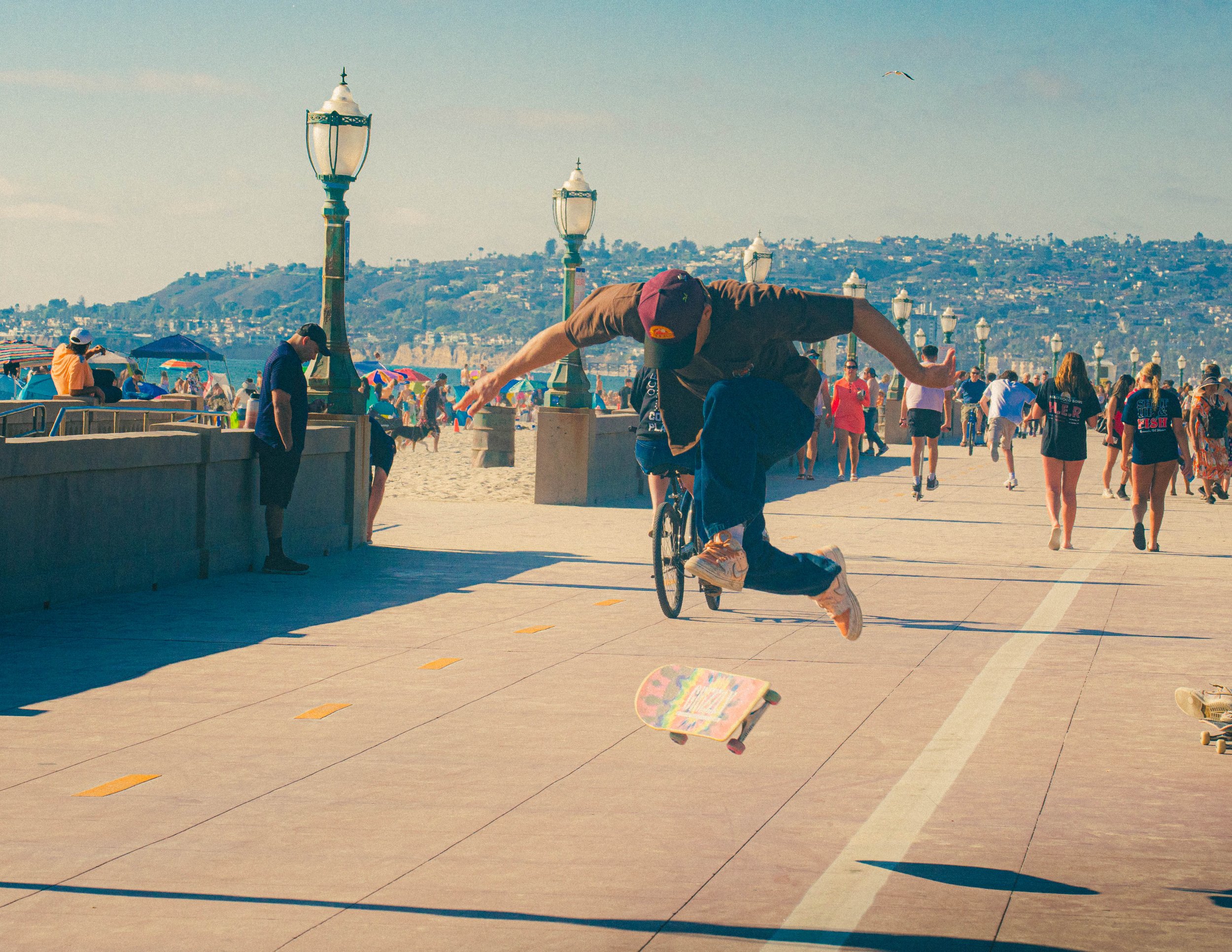 A man on a bicycle in mid-air performing a trick, in front of a crowd on a sunny beach boardwalk with colorful umbrellas, street lamps, and hills in the background.