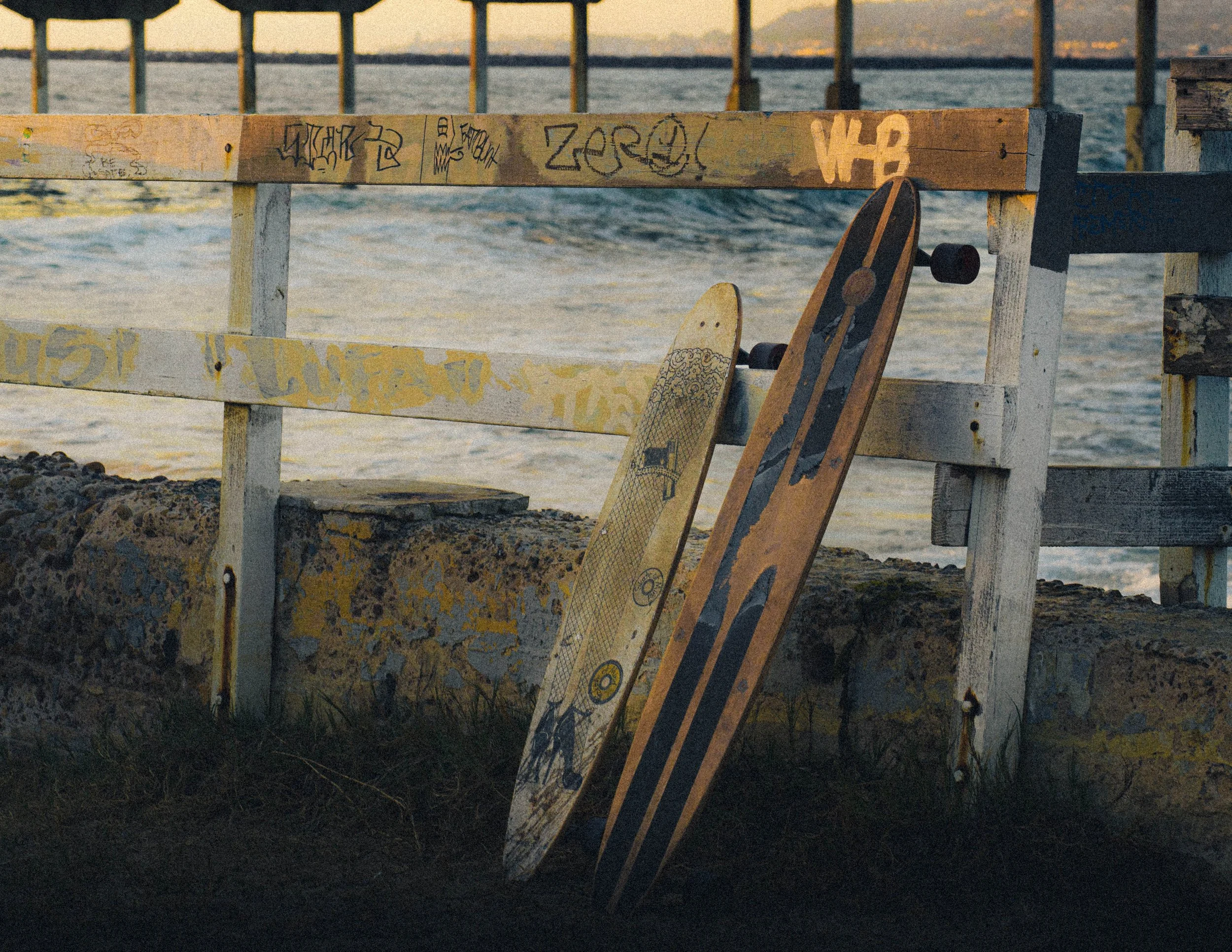 Skateboards leaning against a weathered wooden railing on a beach, with water and a pier in the background during sunset.