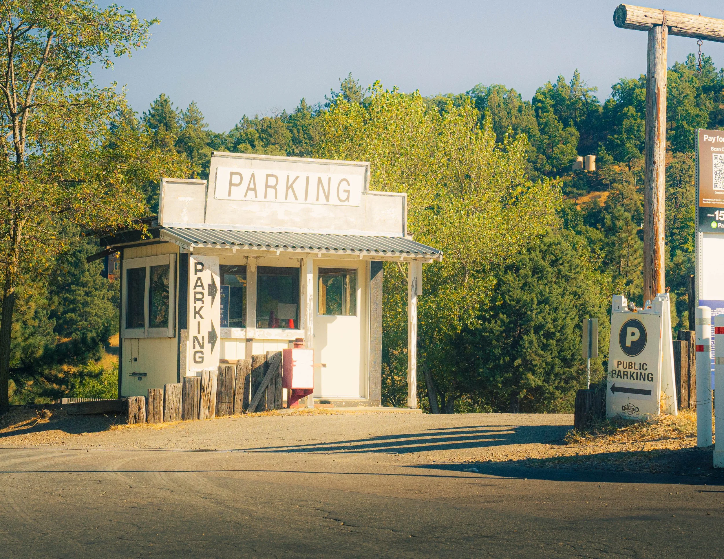 A parking lot entrance with a white building that has a sign reading 'PARKING'. There are trees, signs, and a wooden structure around the area, with surrounding greenery and clear sky in the background.