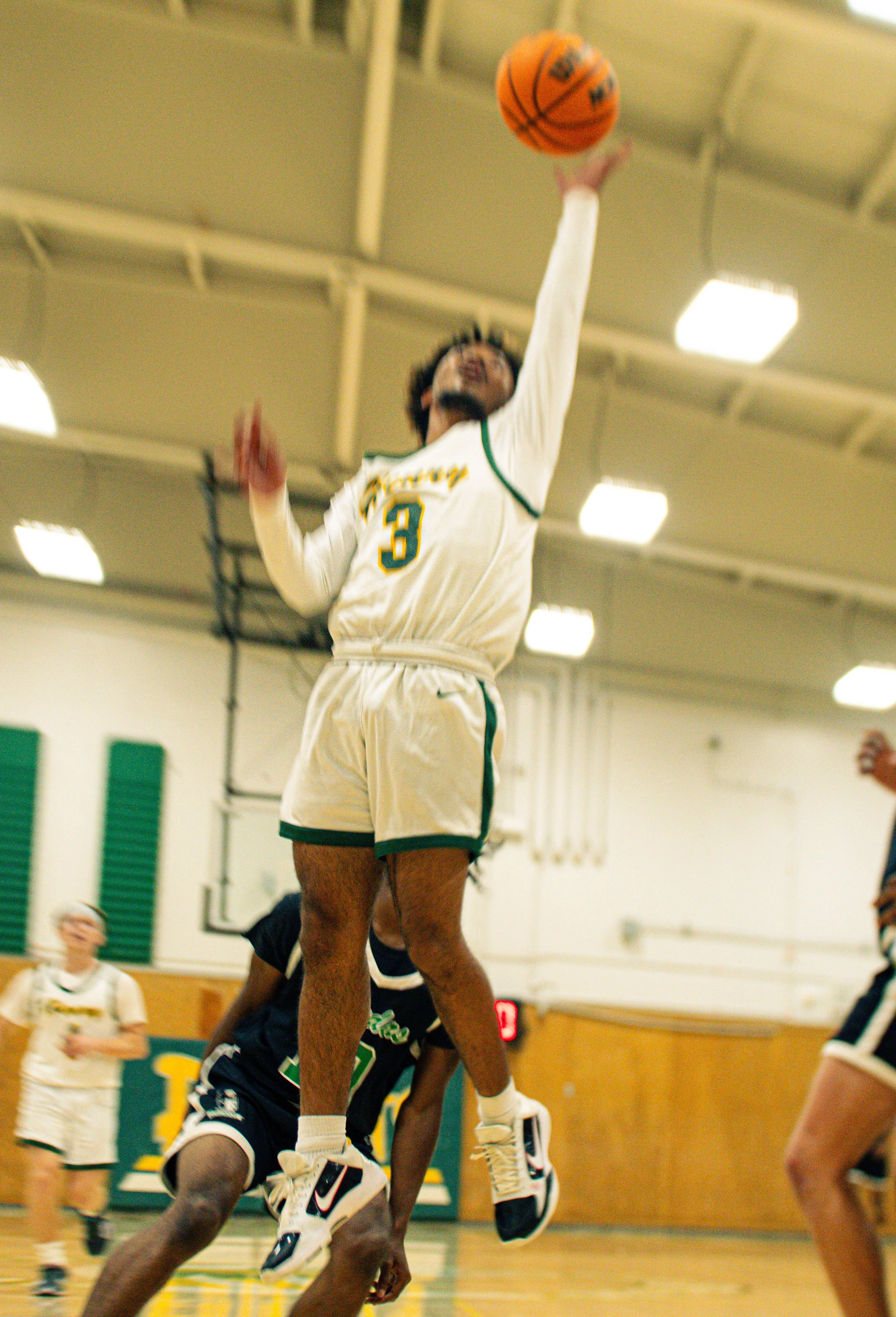 A basketball player in a white jersey with green and yellow accents, number 3, jumping to make a shot during a game in a gymnasium. Other players and a referee are visible in the background.