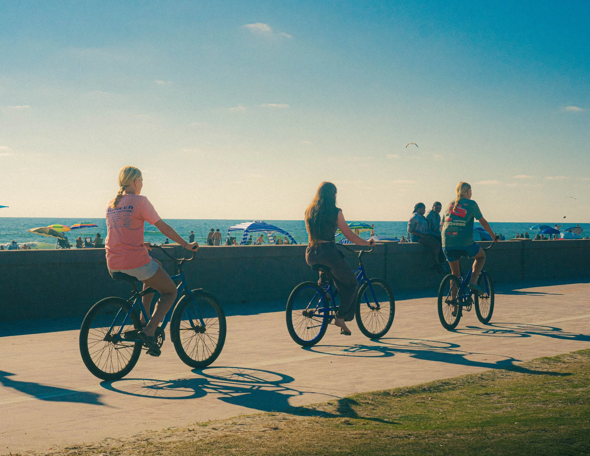 People riding bicycles along a seaside promenade on a sunny day with umbrellas and beachgoers in the background.