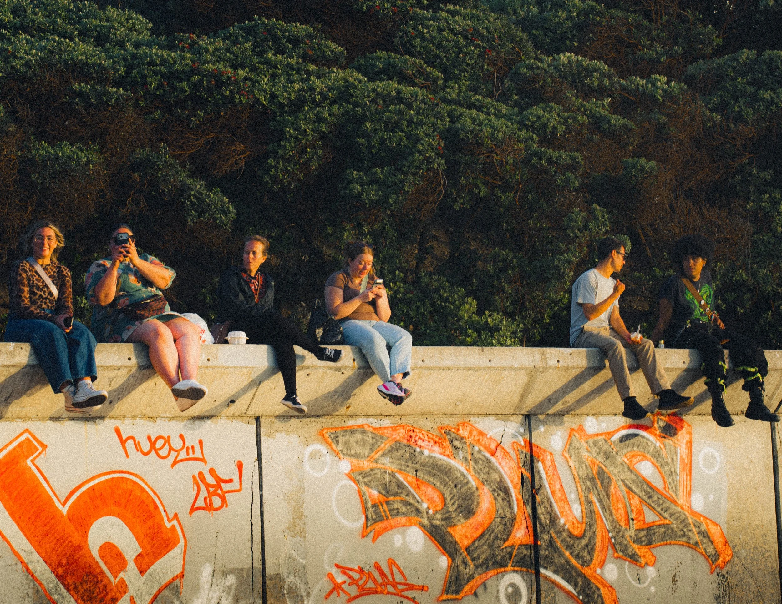 People sitting on a concrete ledge with graffiti, trees in the background, some looking at their phones, others engaging.
