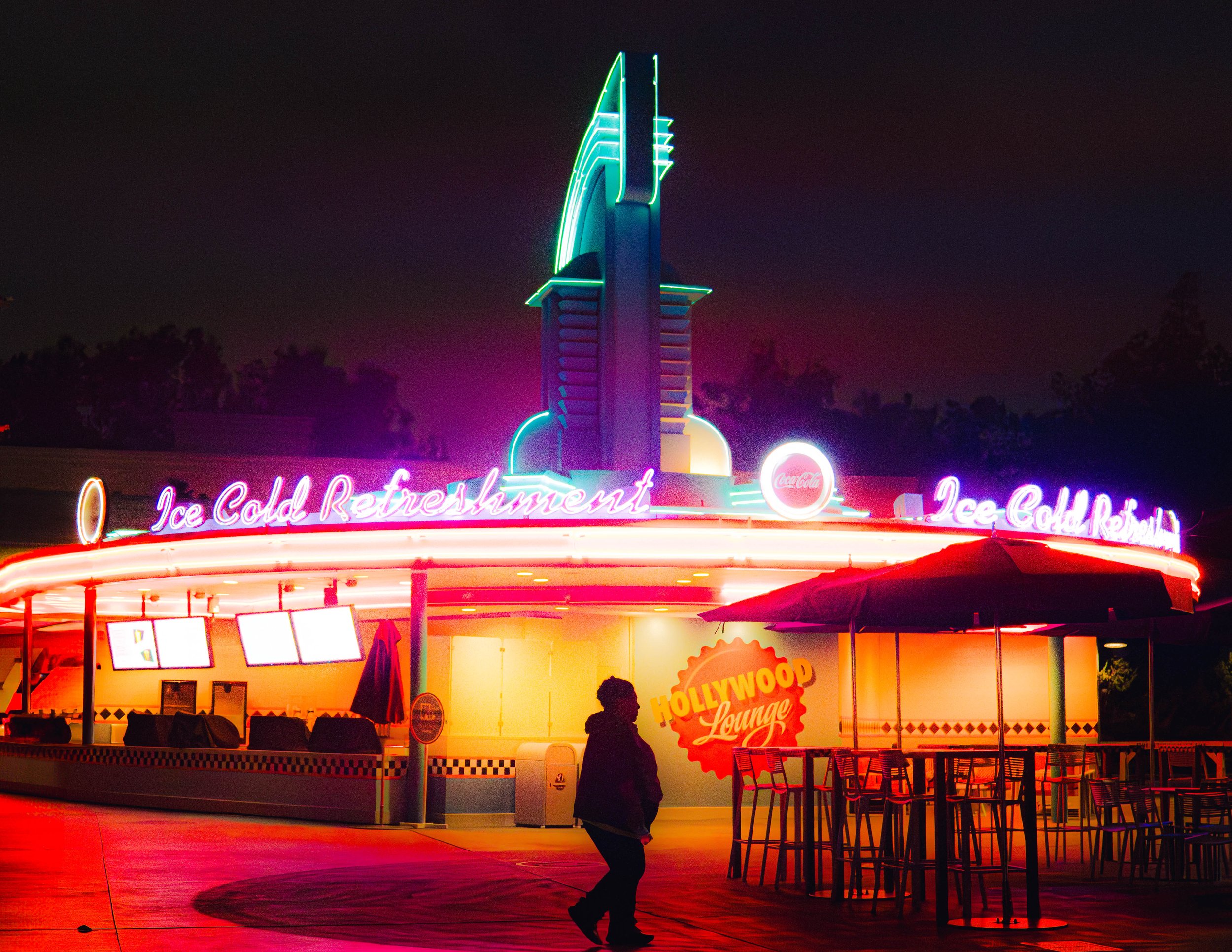 Night view of a retro-style food stand or diner with neon signs advertising 'Ice Cold Refreshment'. Tall neon sign structure behind the stand, with pink, green, and blue lighting, creating a vintage Hollywood lounge atmosphere. Silhouettes of people 