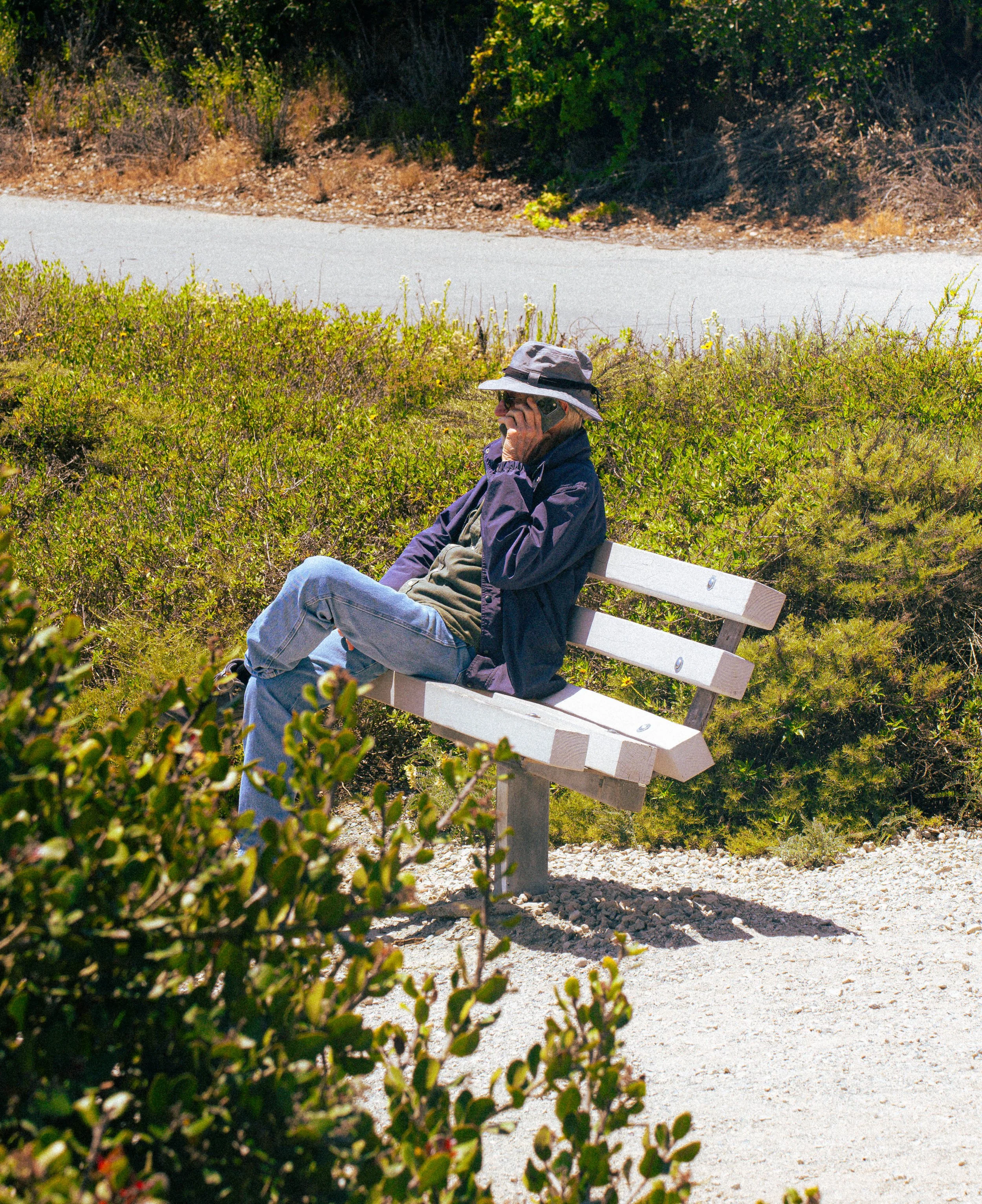 A man wearing a wide-brim hat and sunglasses is sitting on a park bench beside lush green bushes, talking on a cellphone on a sunny day.