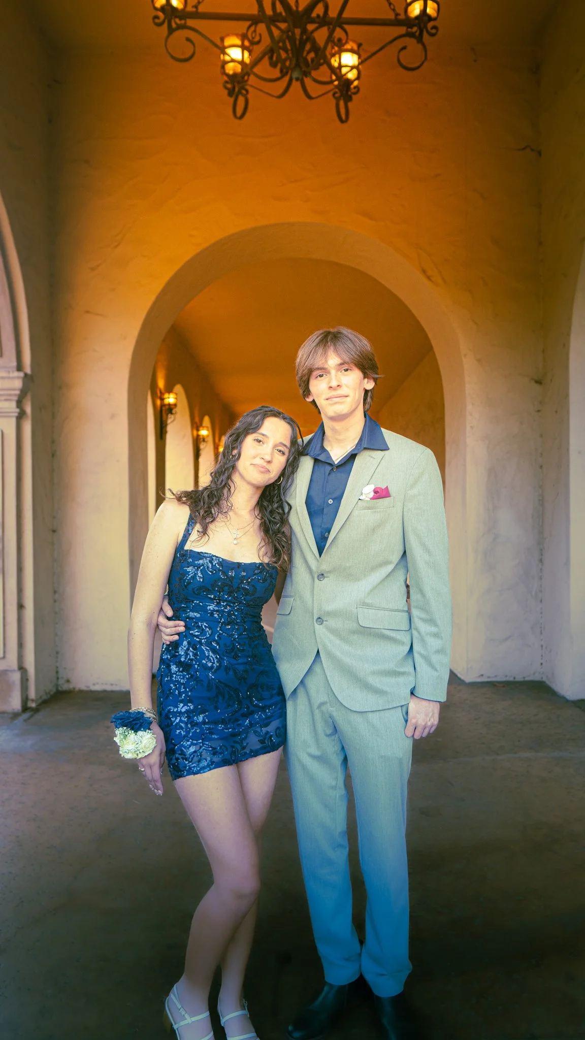 A young woman in a blue lace dress and a young man in a light gray suit stand together in an arched corridor under a chandelier, smiling for a photo.