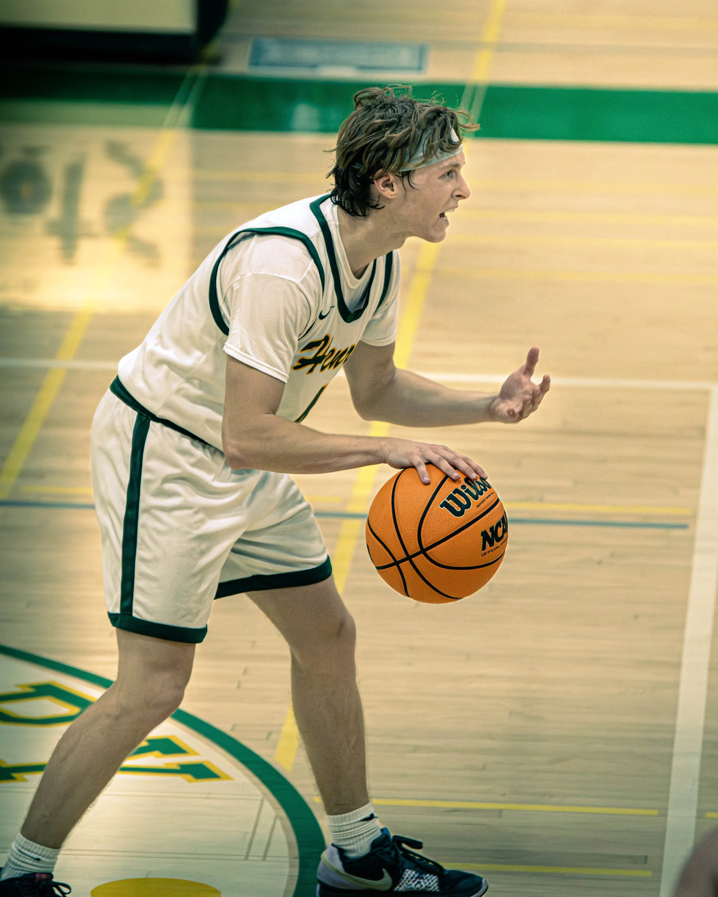 A basketball player in a white and green uniform, wearing a headband, dribbling a Wilson basketball on a gymnasium court.