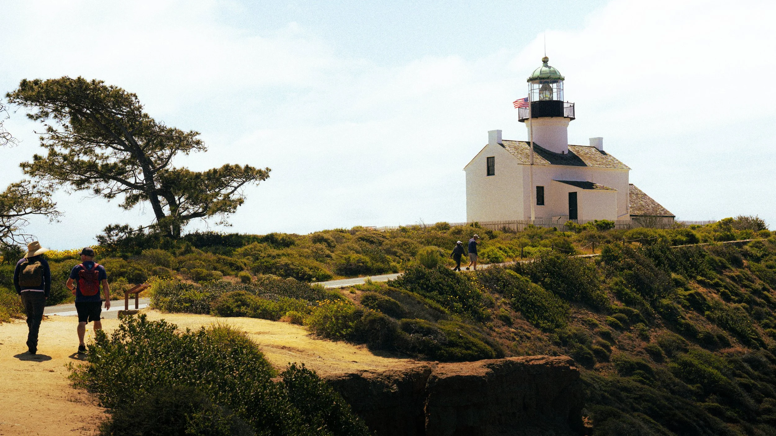 People hiking on a trail near a white lighthouse with a green dome and an American flag, surrounded by shrubbery and a few trees, under a cloudy sky.