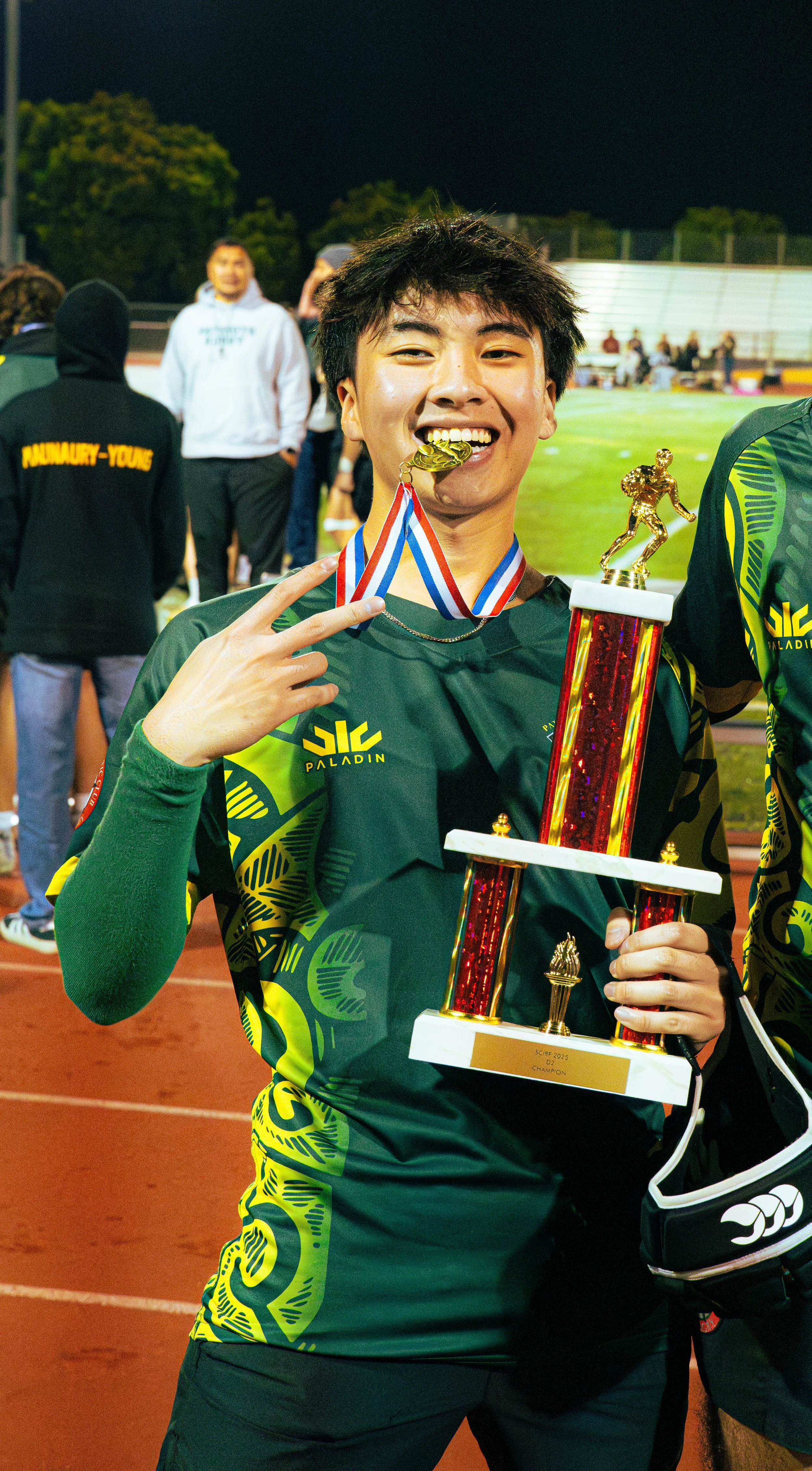 Young male athlete in a green and black sports uniform holding a trophy, smiling, and making a peace sign with his right hand on a sports field at night.