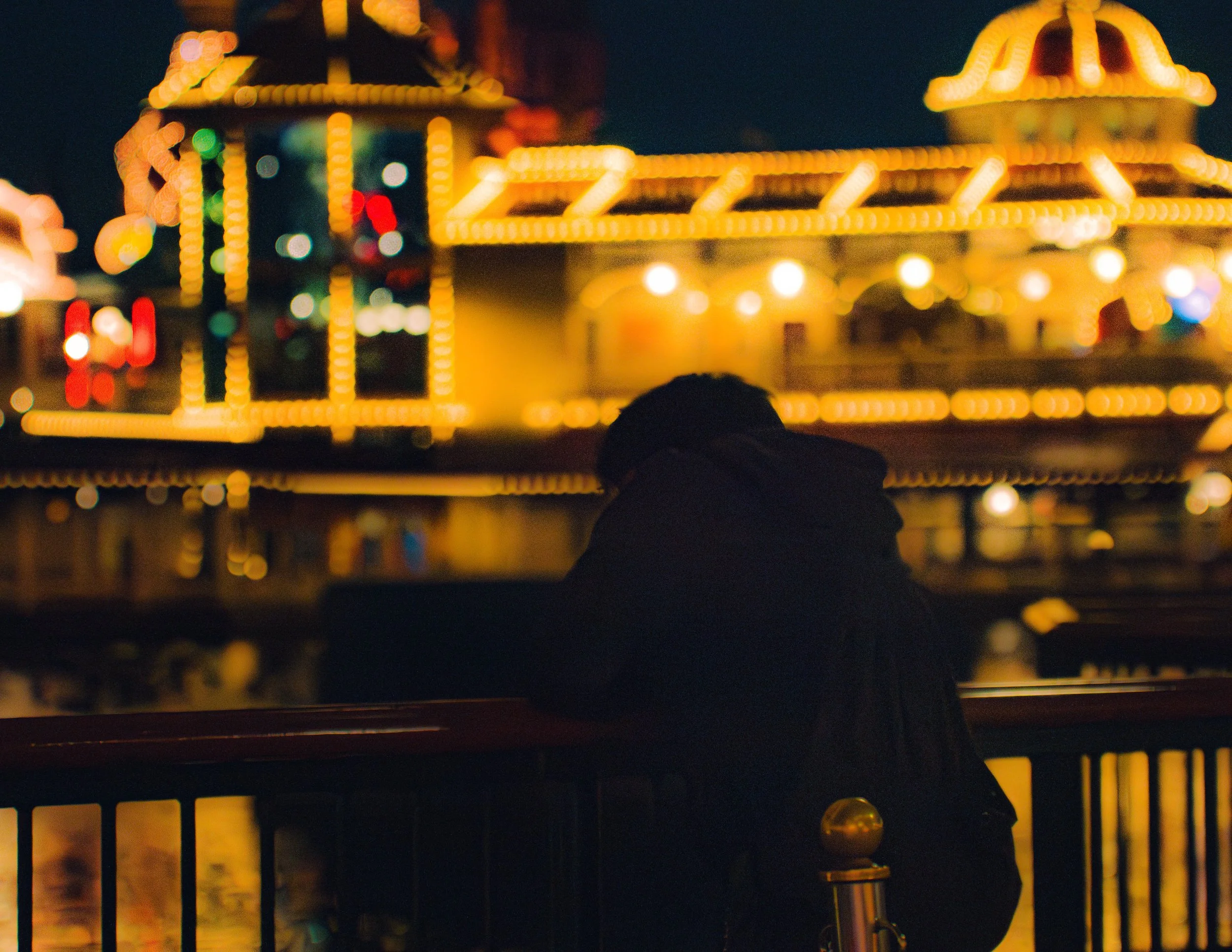 Nighttime view of a brightly lit amusement park ride with yellow lights, featuring a castle or tower structure and a connecting bridge, with a silhouette of a person's head in the foreground.