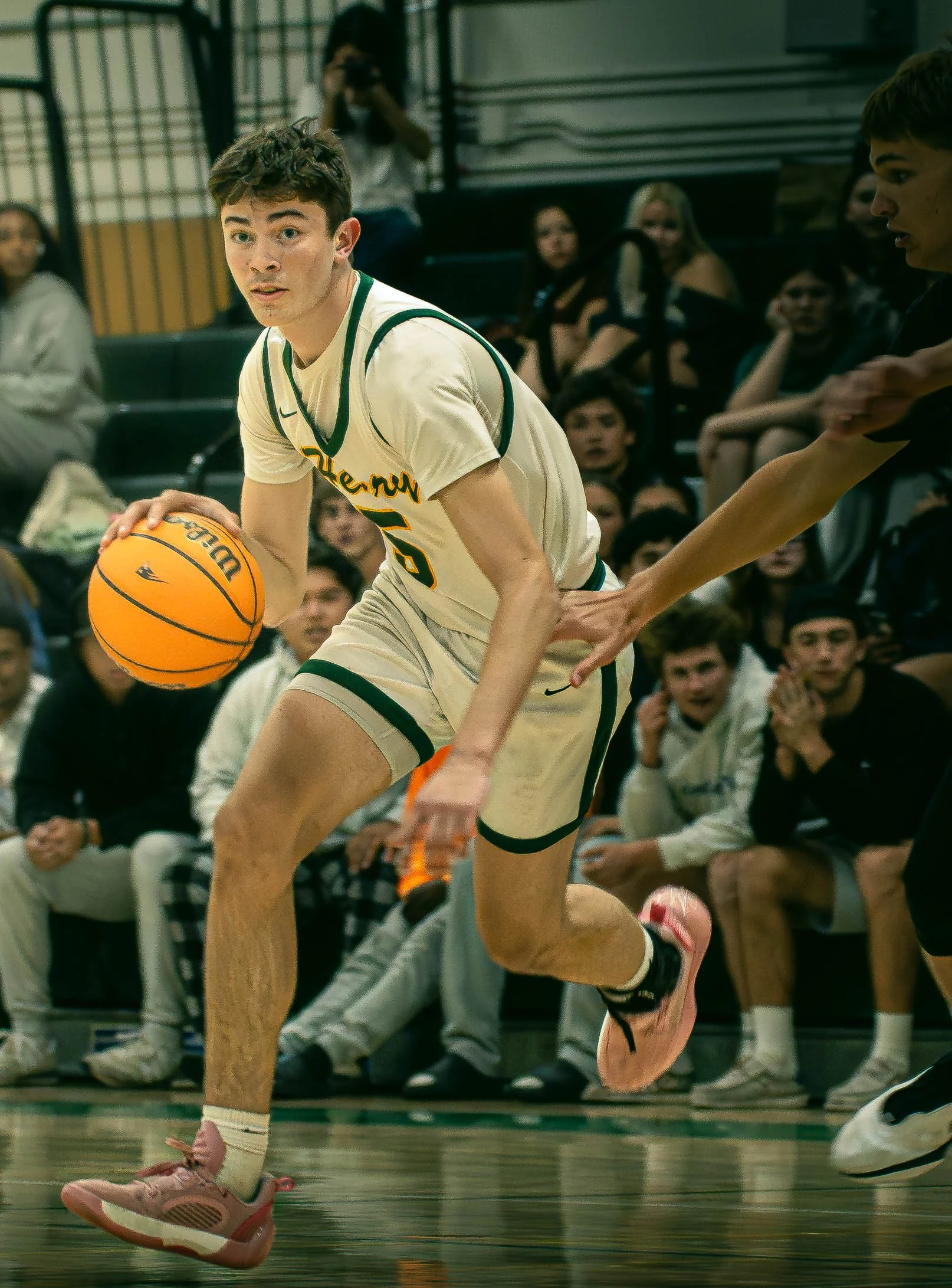 A basketball game in progress with a player dribbling the ball, wearing a white jersey with green accents, while another player reaches for the ball. Audience members are seen sitting on benches in the background.
