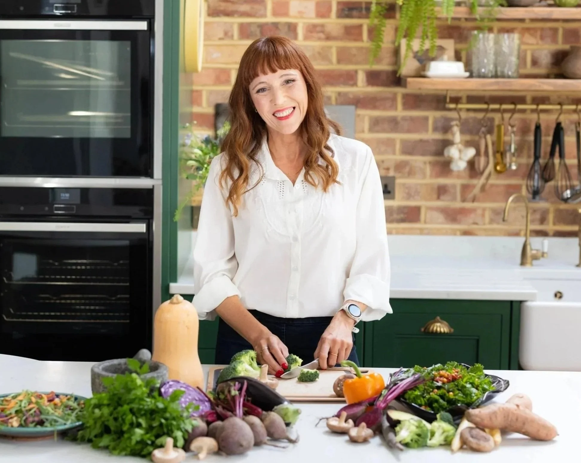 A woman smiling in a kitchen, chopping vegetables on a white countertop surrounded by various fresh greens, vegetables and fruit.