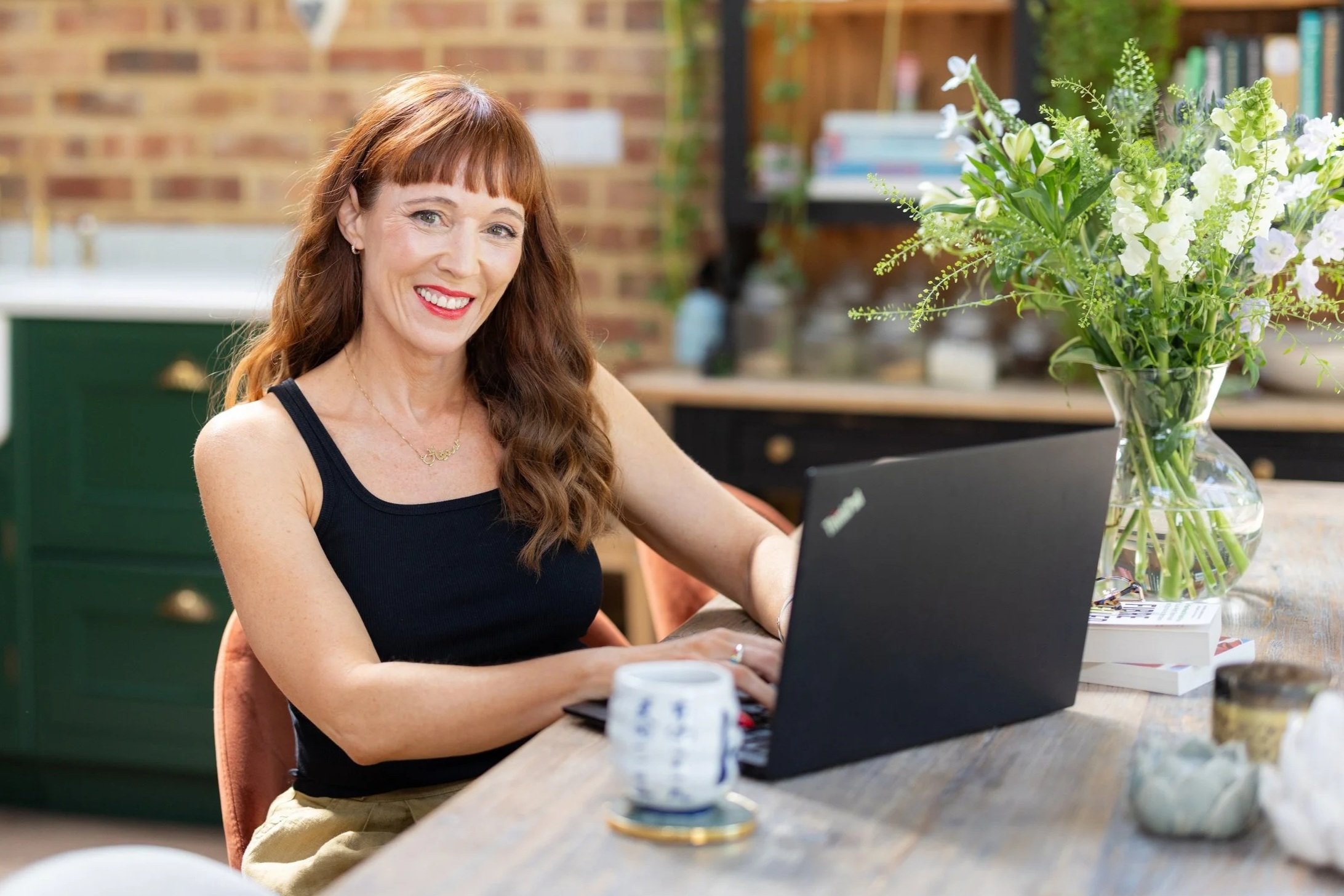 A woman is smiling while working on a laptop at a wooden table, with a large bouquet of white and green flowers in a glass vase nearby.