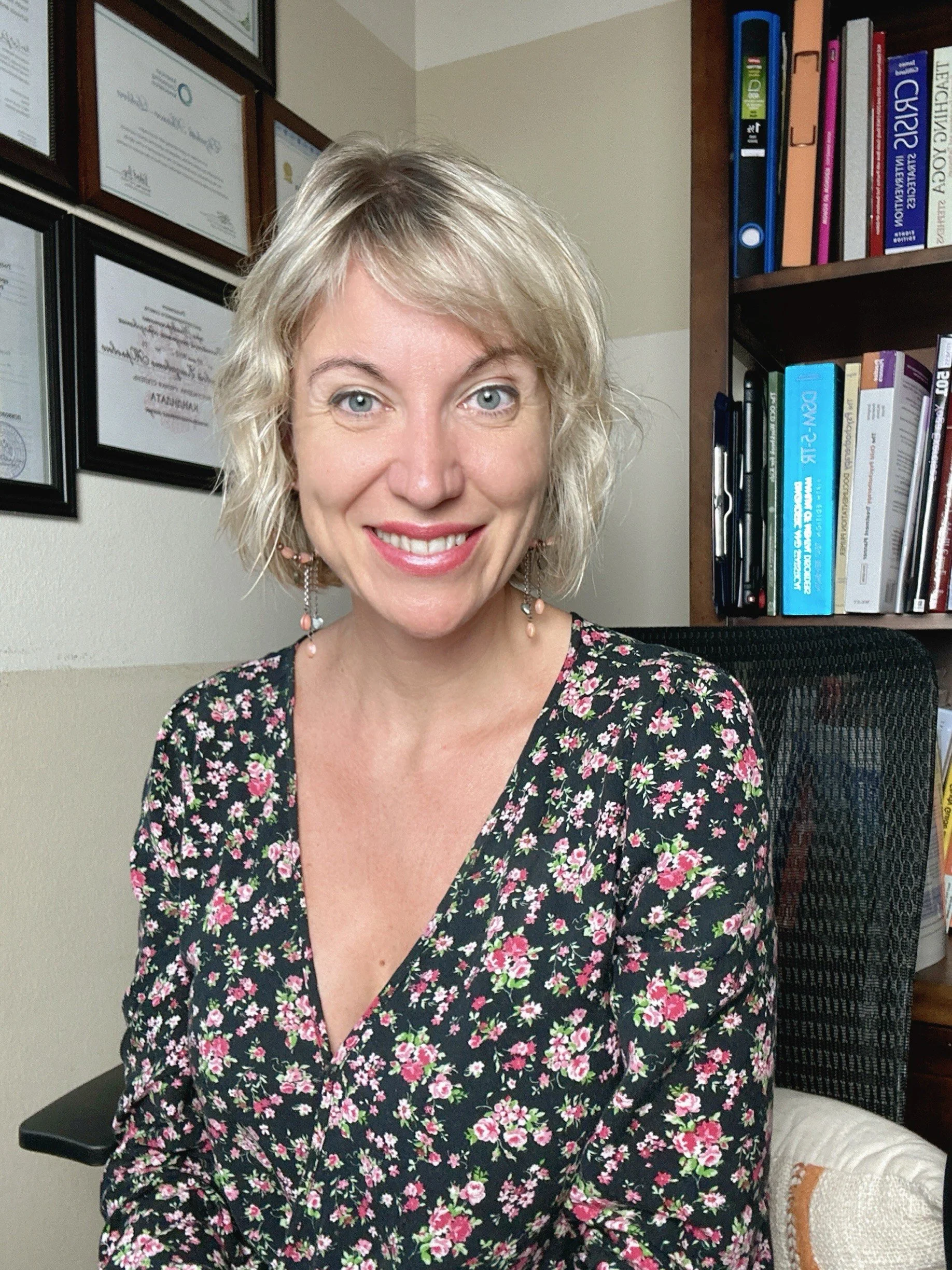 A woman with blonde, wavy hair and light blue eyes smiling, wearing a floral dress with pink, white, and green flowers on a dark background, sitting in an office with framed certificates on the wall and a bookshelf filled with binders and books nearby.