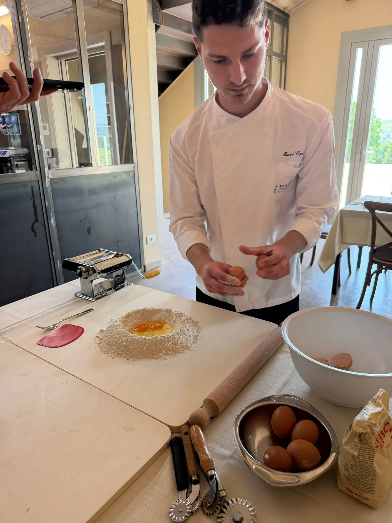 A man preparing to make pasta dough with eggs on a floured surface, surrounded by kitchen tools, eggs in a metal bowl, a rolling pin, and a bag of flour, in a well-lit kitchen with a window view outside.