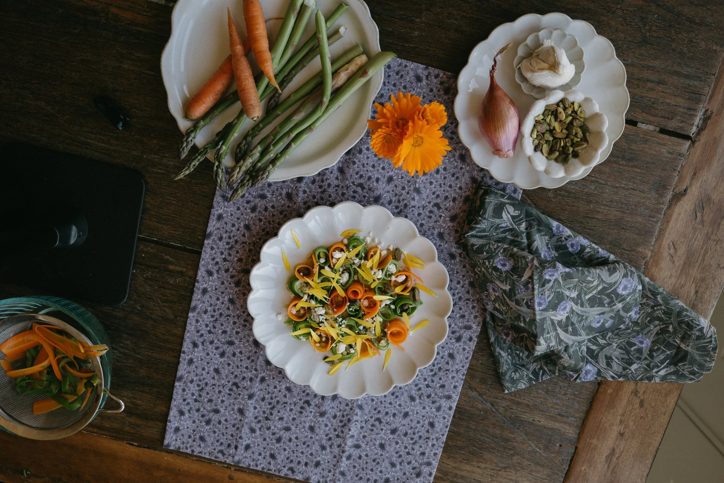A top view of a rustic wooden table with a purple patterned tablecloth, showcasing various fresh vegetables, flowers, and prepared salads on white dishes.