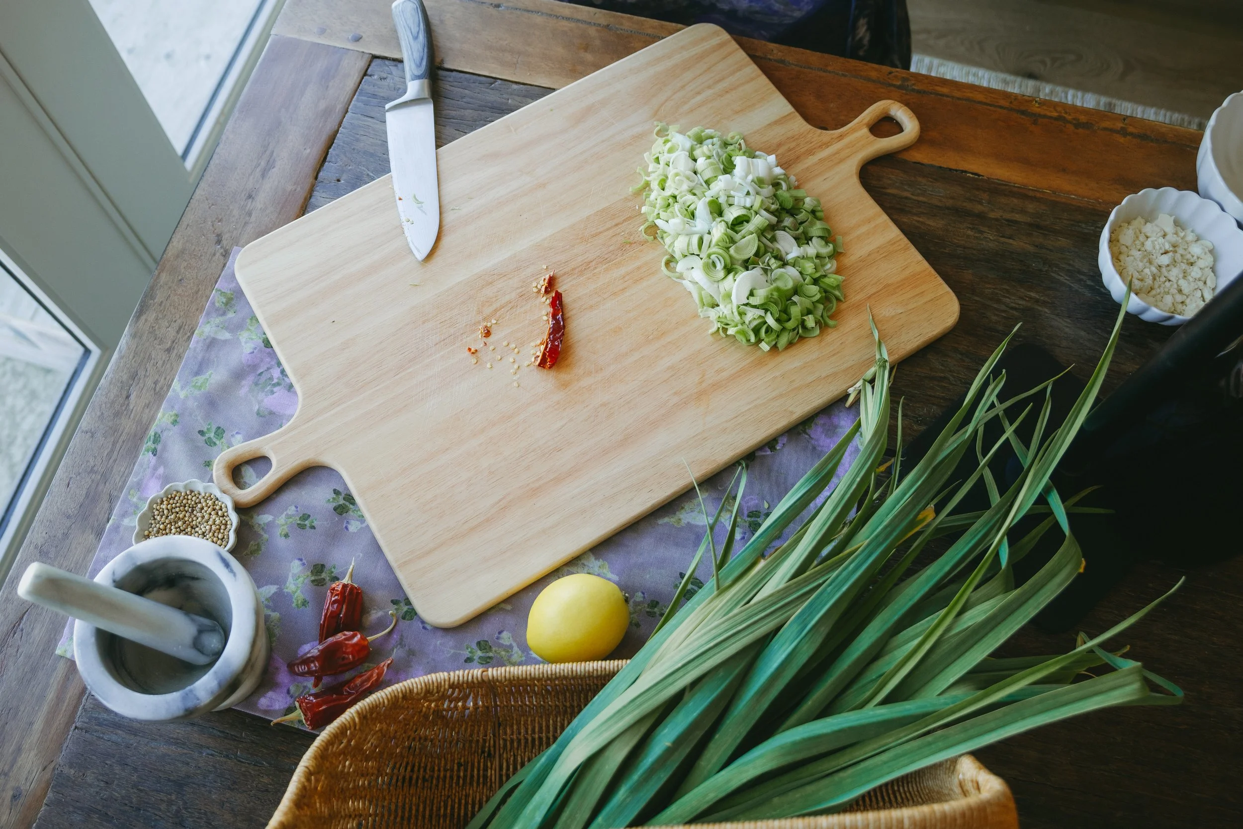 A wooden cutting board with chopped green onions, a knife, dried red chili peppers, and bowls of crumbled cheese and peppercorns on a rustic wooden table near a window.