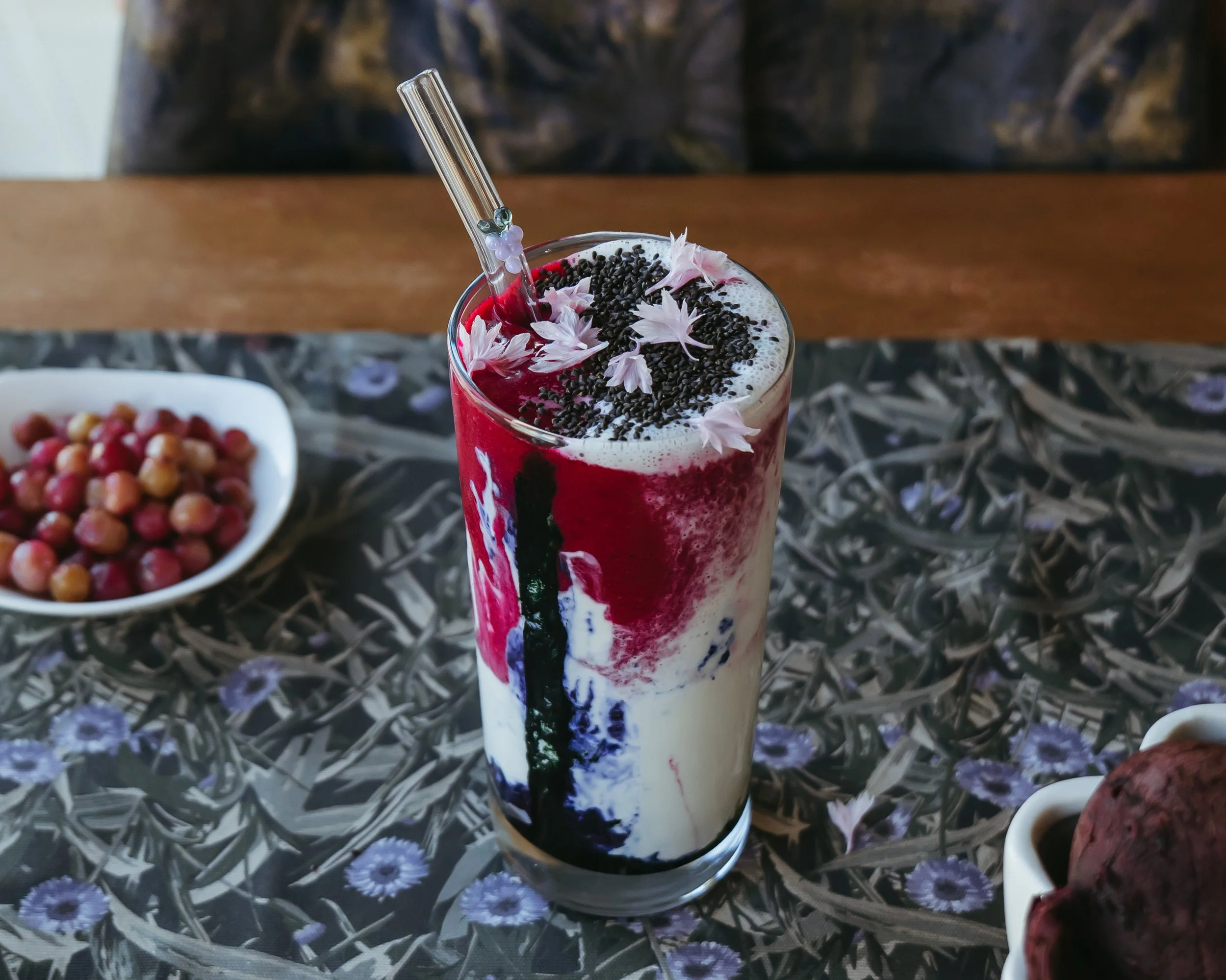 A colorful drink topped with black sprinkles and pink flower petals, served with a clear straw, on a decorative table with a bowl of fruit in the background.