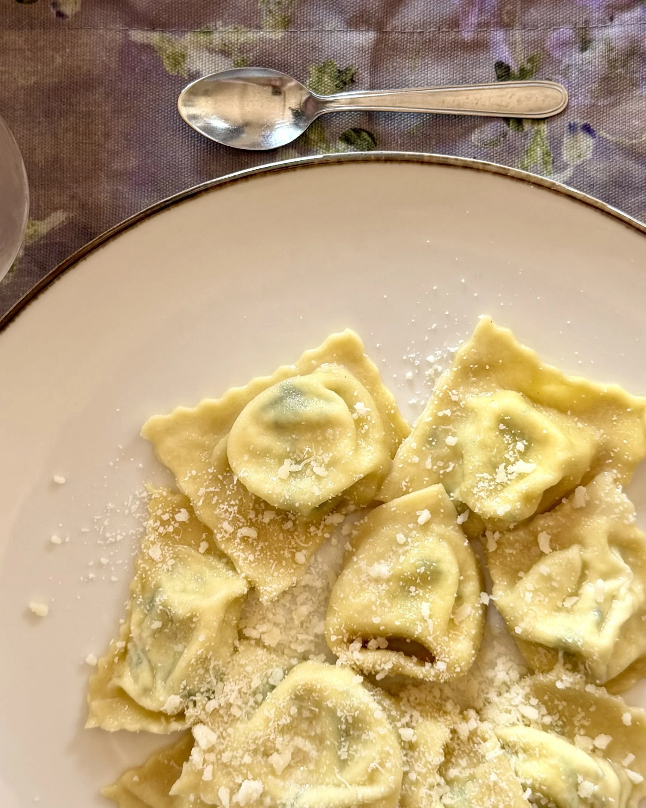 Close-up of a white plate filled with cooked tortellini pasta sprinkled with grated cheese, on a floral-patterned tablecloth, with a silver spoon placed above the plate.